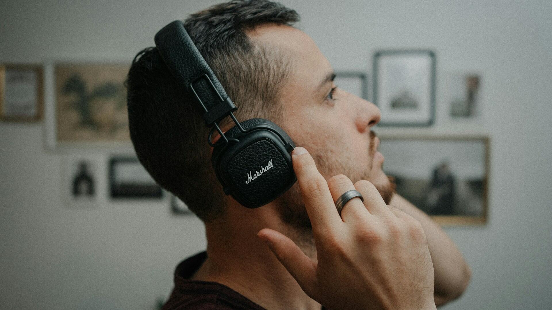 A person wearing black Marshall headphones gestures with their hand, surrounded by framed pictures on the wall.