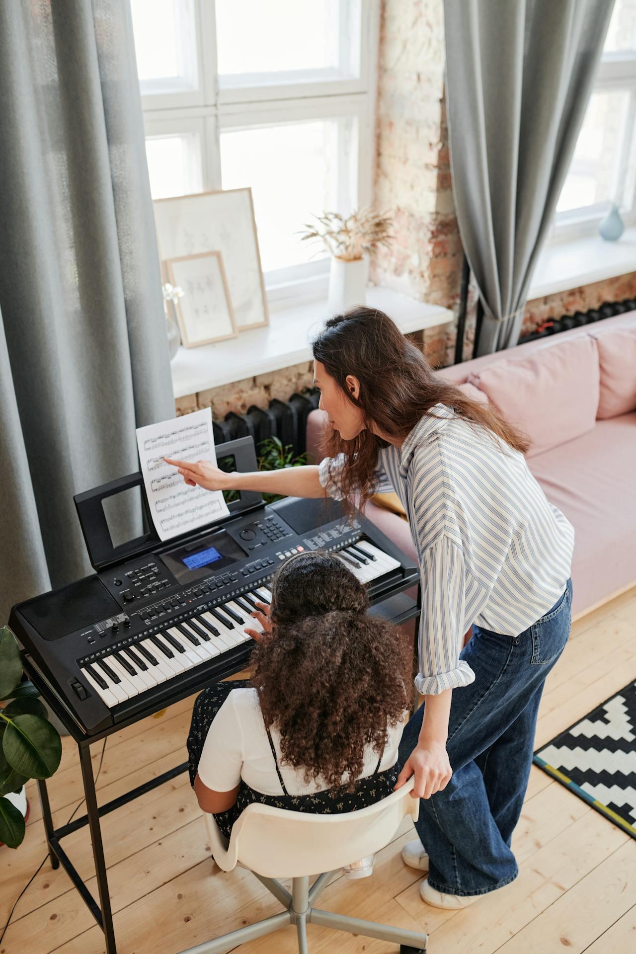 A woman teaches a girl at a keyboard, pointing at sheet music, in a cozy room with large windows and a pink couch.