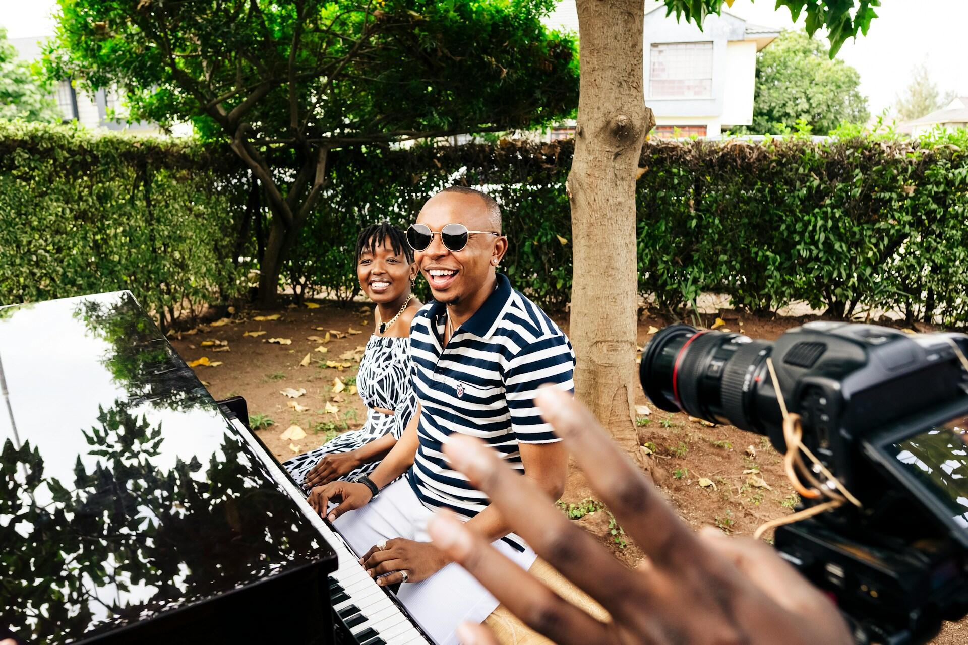 A man in a striped shirt and a woman in a patterned dress sit at a piano, surrounded by lush greenery, as someone records the moment.