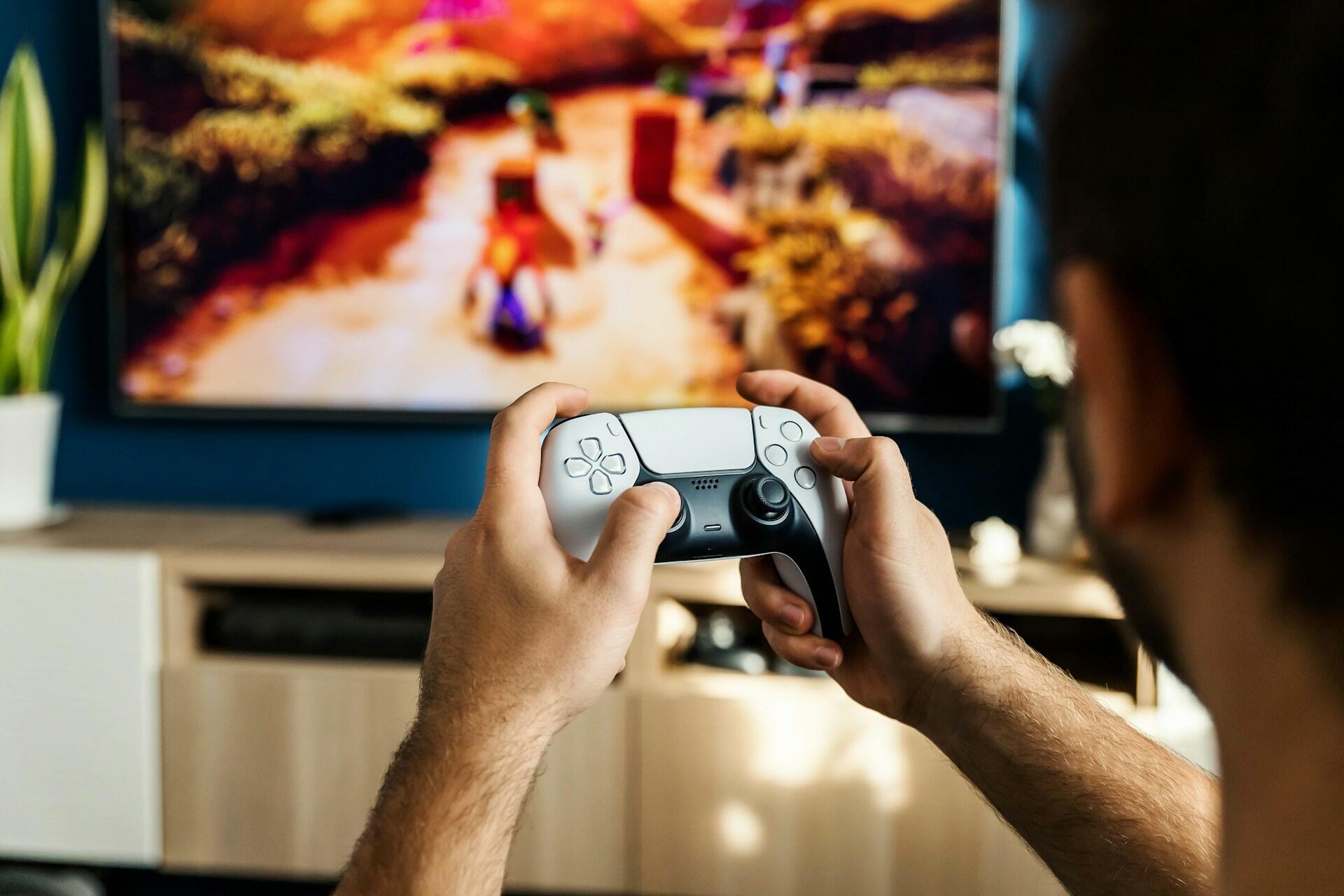 A person holds a PS5 controller in front of a TV displaying a vibrant video game landscape. A potted plant is visible nearby.