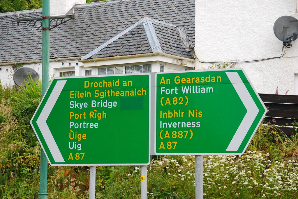 Green road signs in Gaelic and English, directing to Skye Bridge, Portree, and Fort William amidst a lush floral backdrop.
