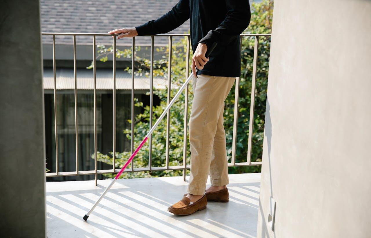 A person with a white cane stands on a balcony, gently resting a hand on the railing, with greenery visible in the background.