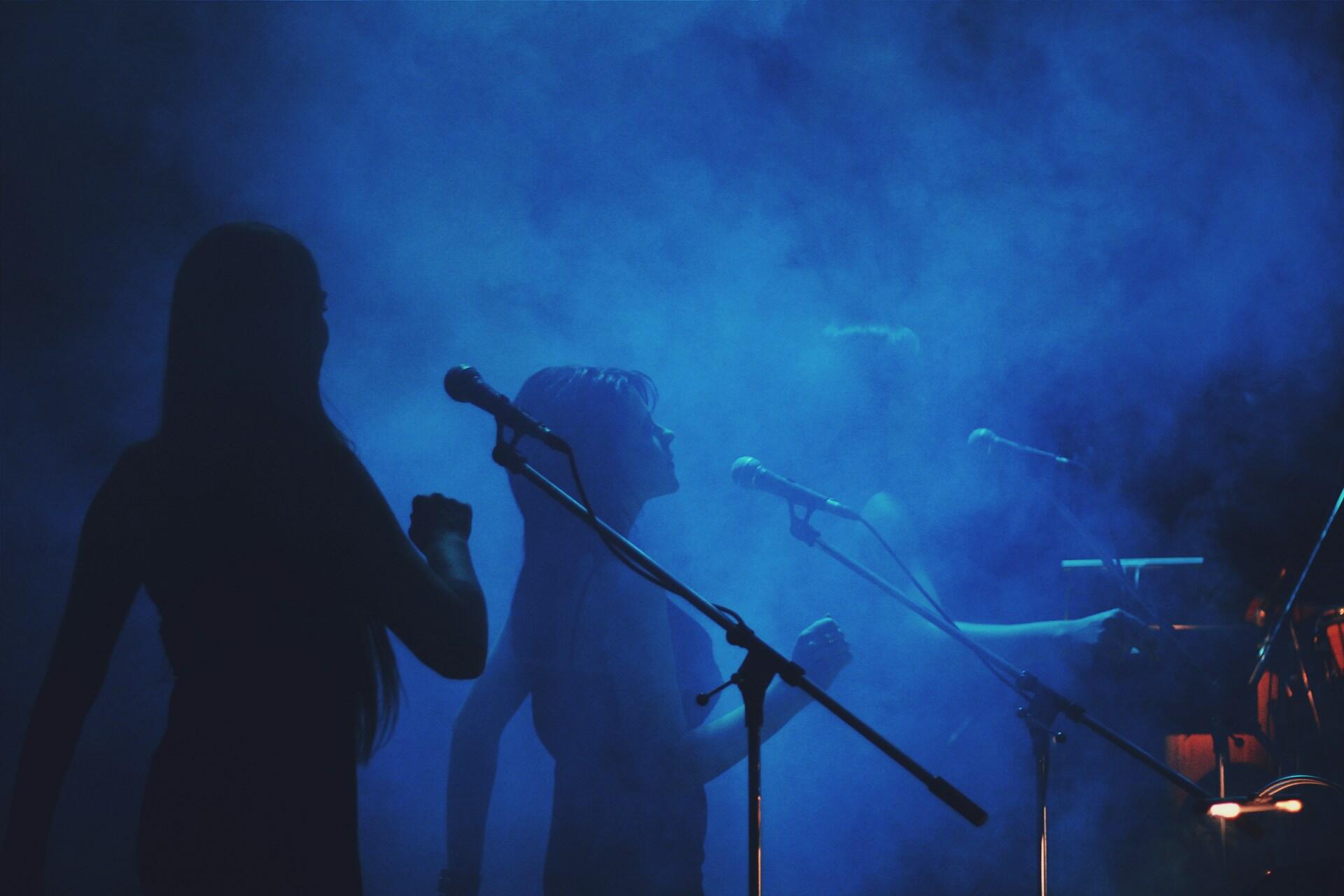 Silhouettes of performers in a smoky, blue-lit atmosphere, with microphones positioned for a live music performance.