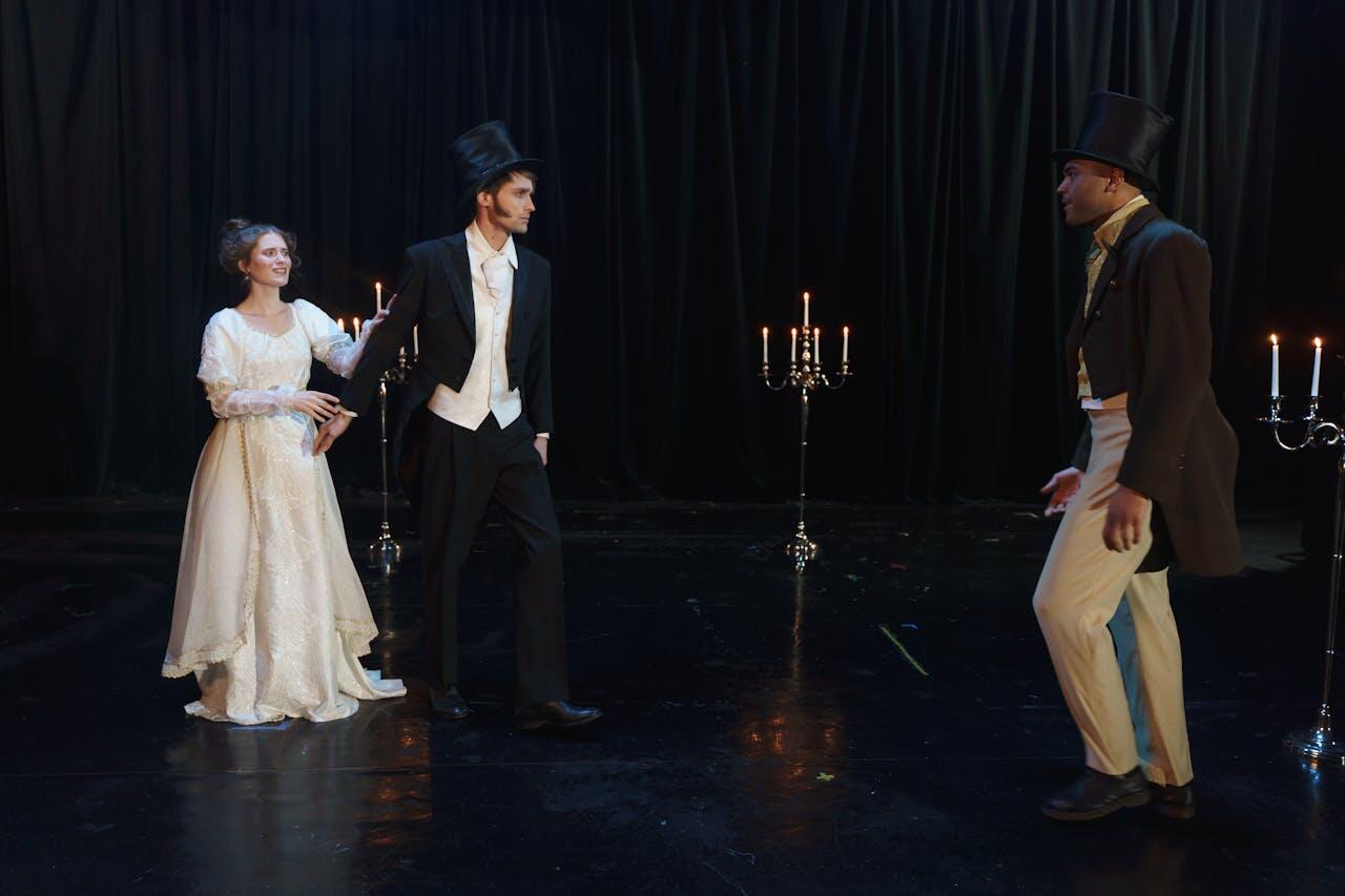Three performers in elegant costumes on stage, with candelabras providing a warm glow against a dark backdrop.