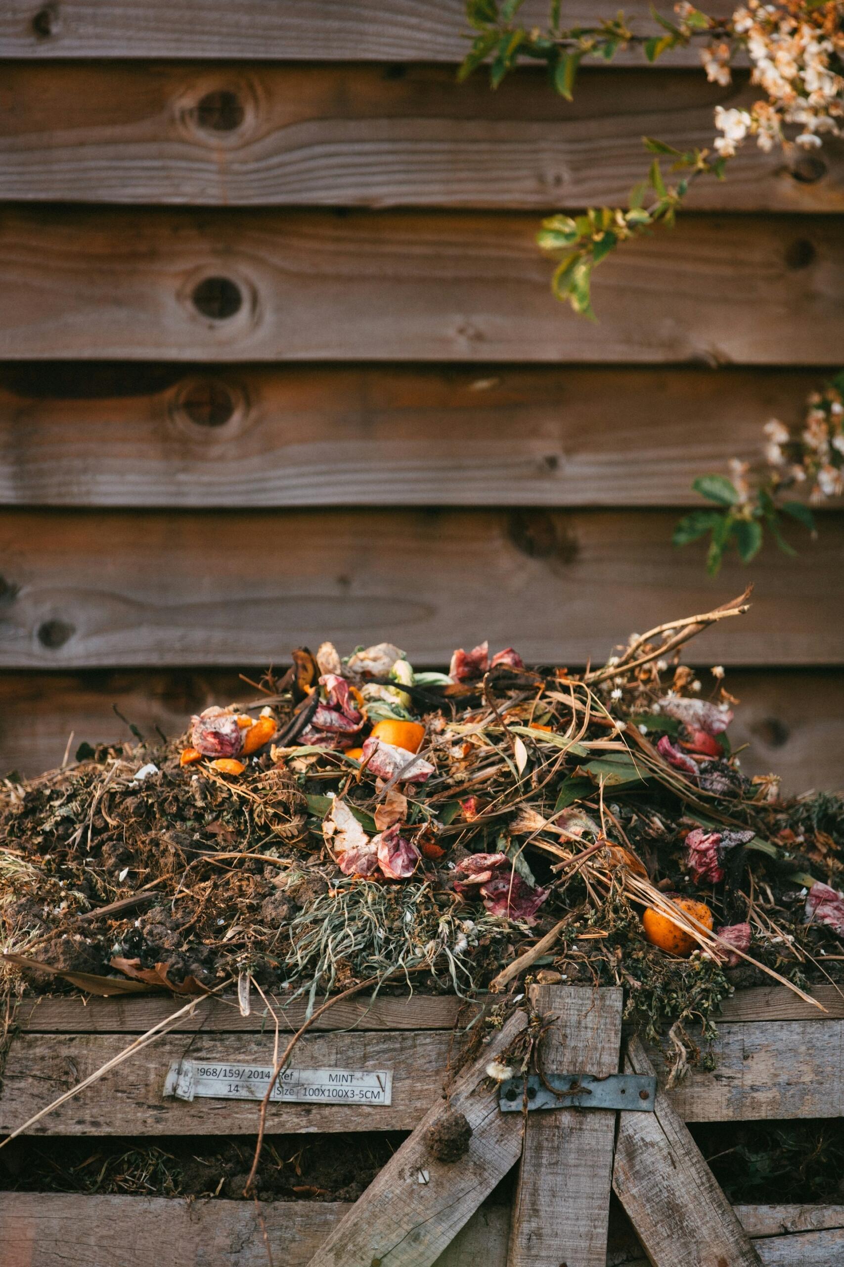 A wooden compost bin overflowing with kitchen scraps, leaves, and twigs, set against a weathered wood background.