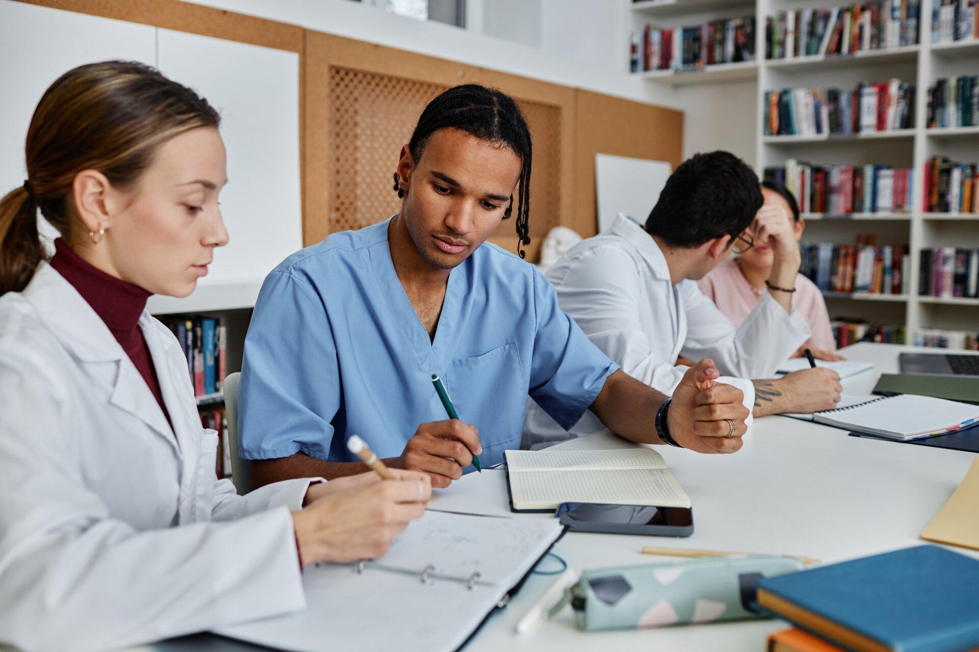 A diverse group of medical students studying together in a library setting, wearing lab coats and scrubs, surrounded by notebooks and textbooks.