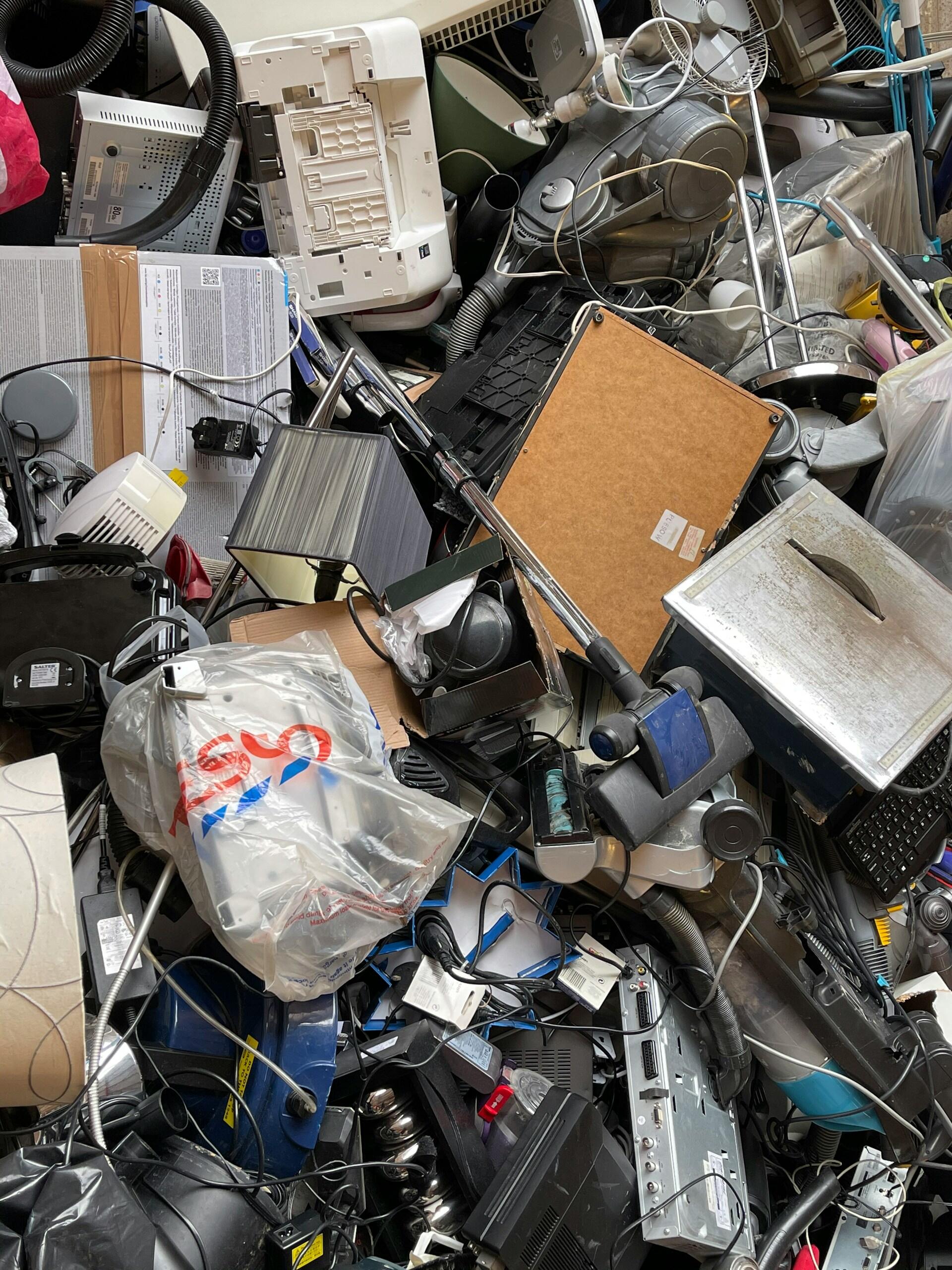 A chaotic heap of discarded electronics, including printers, cables, appliances, and various small devices, piled high.