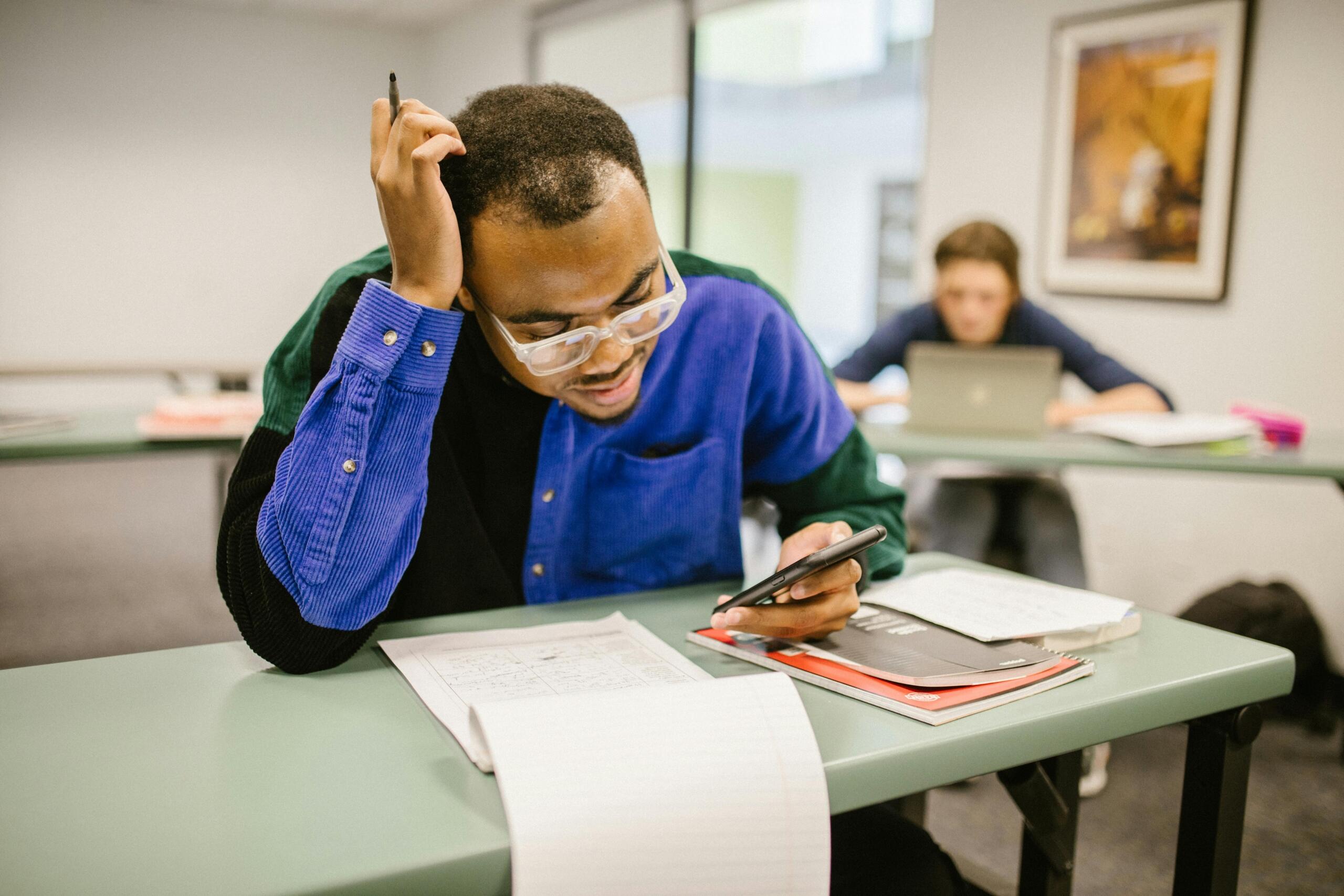 Student studying for exams at a classroom desk while reviewing notes and using a smartphone