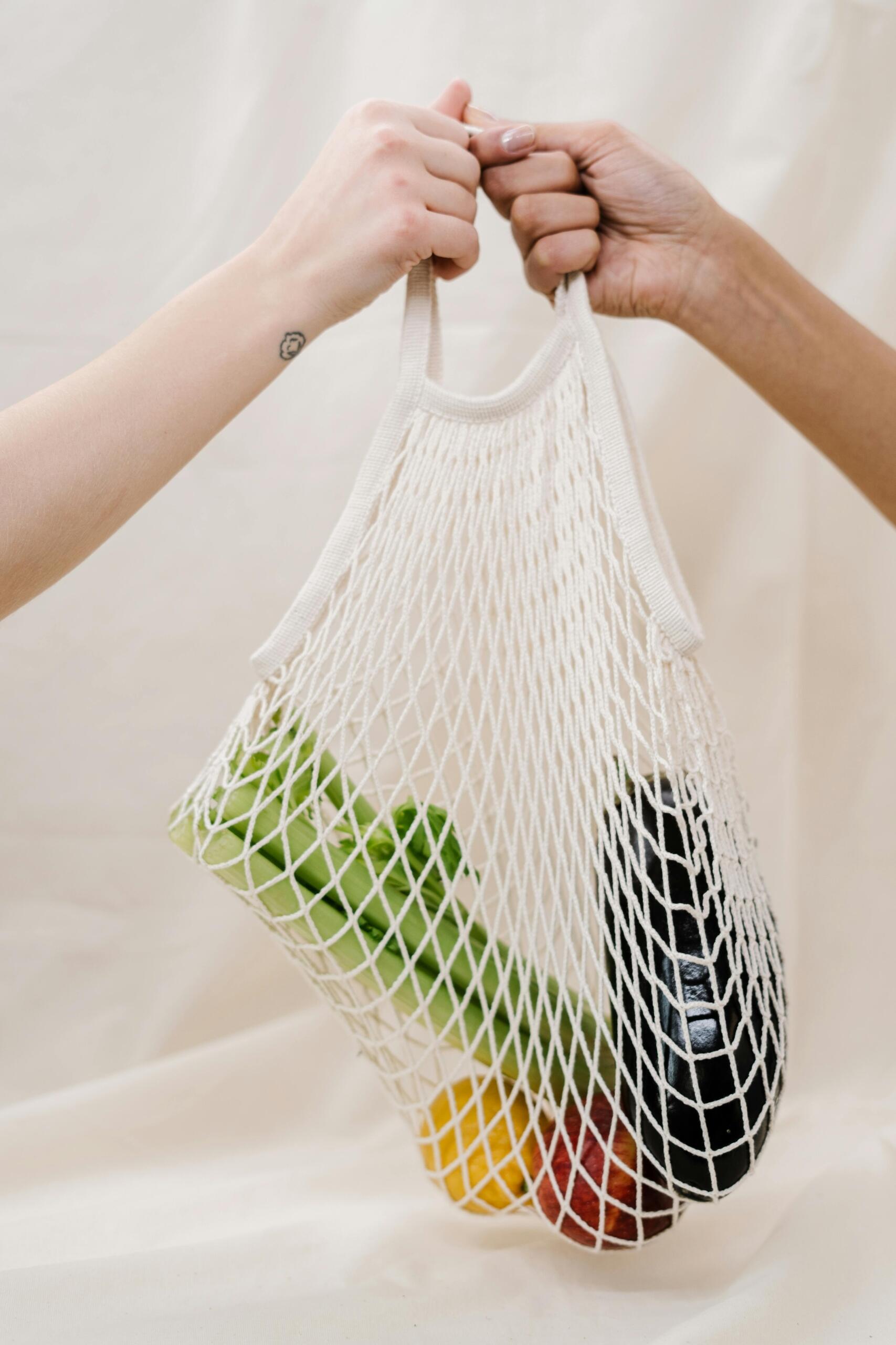 Two people holding a reusable net grocery bag filled with fresh vegetables and fruits.
