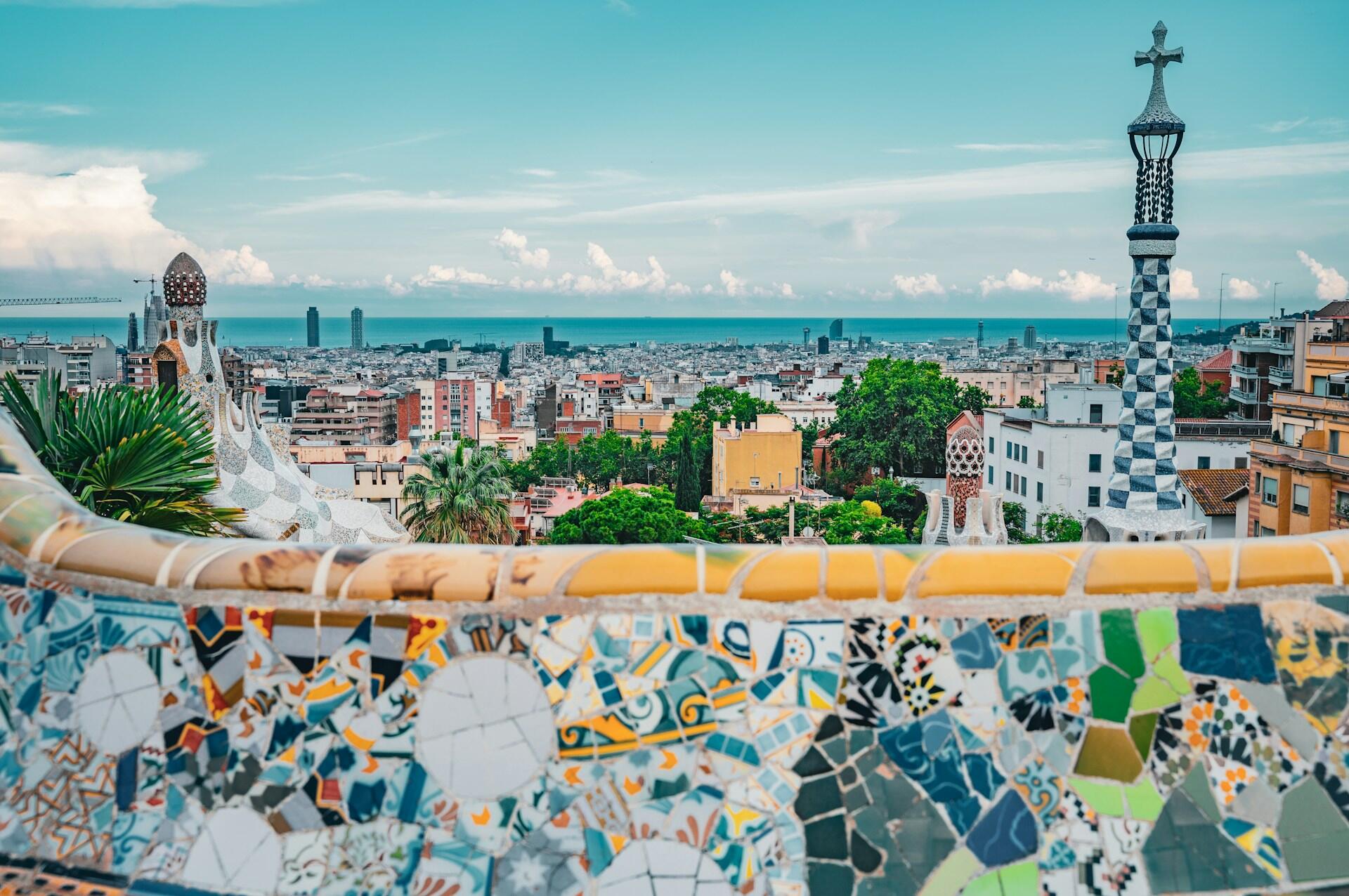 Panoramic view of Barcelona from Park Güell, showcasing colorful mosaics, city buildings, and the sea under a clear blue sky.