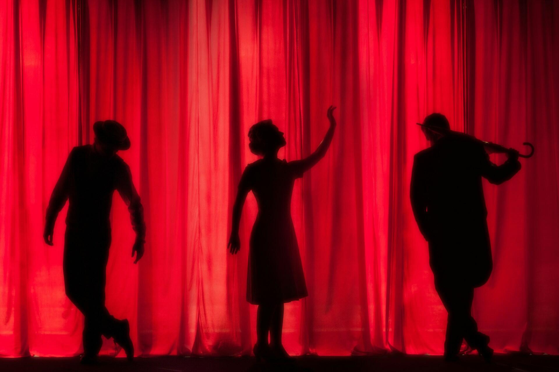 Silhouettes of three performers against a vibrant red curtain, striking poses in an elegant theatre setting.