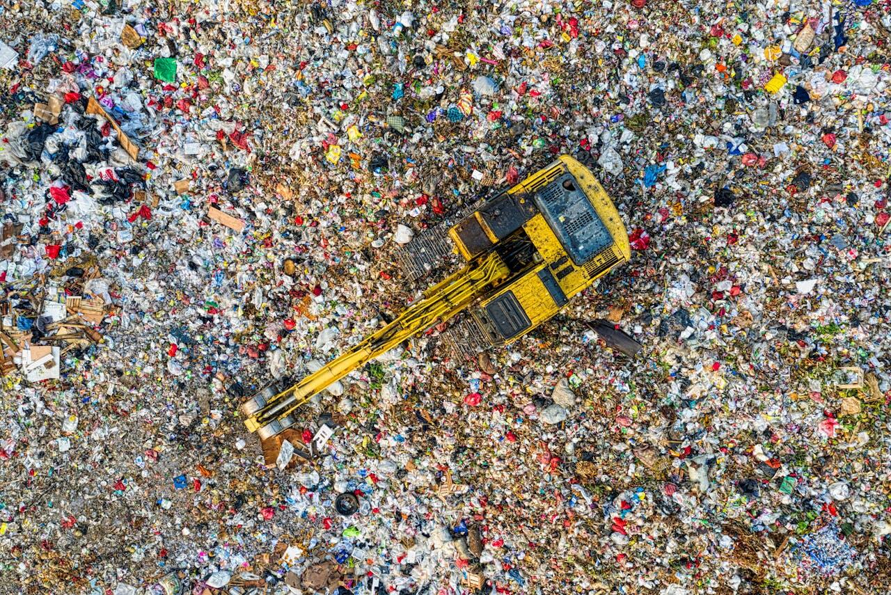 Aerial view of a yellow excavator atop a sprawling landfill, surrounded by extensive litter and waste materials.