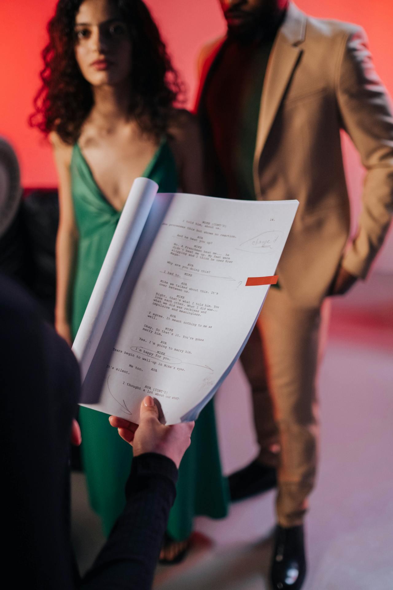 A person holds a script with dialogue, while two actors in stylish outfits stand nearby against a vibrant red backdrop.