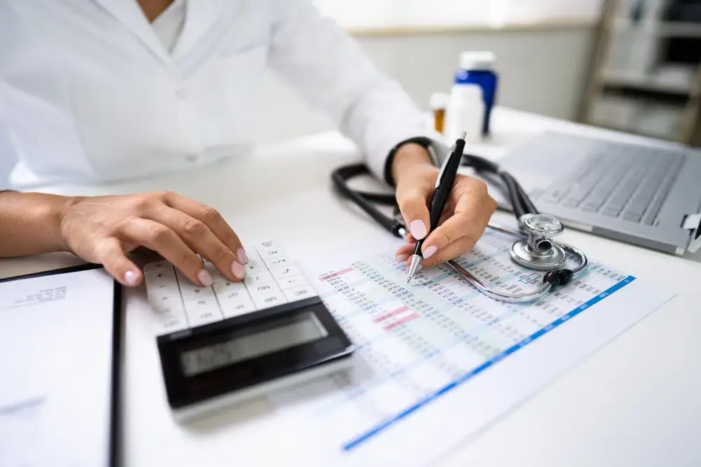 Medical student calculating expenses with a stethoscope, laptop, and financial documents on desk