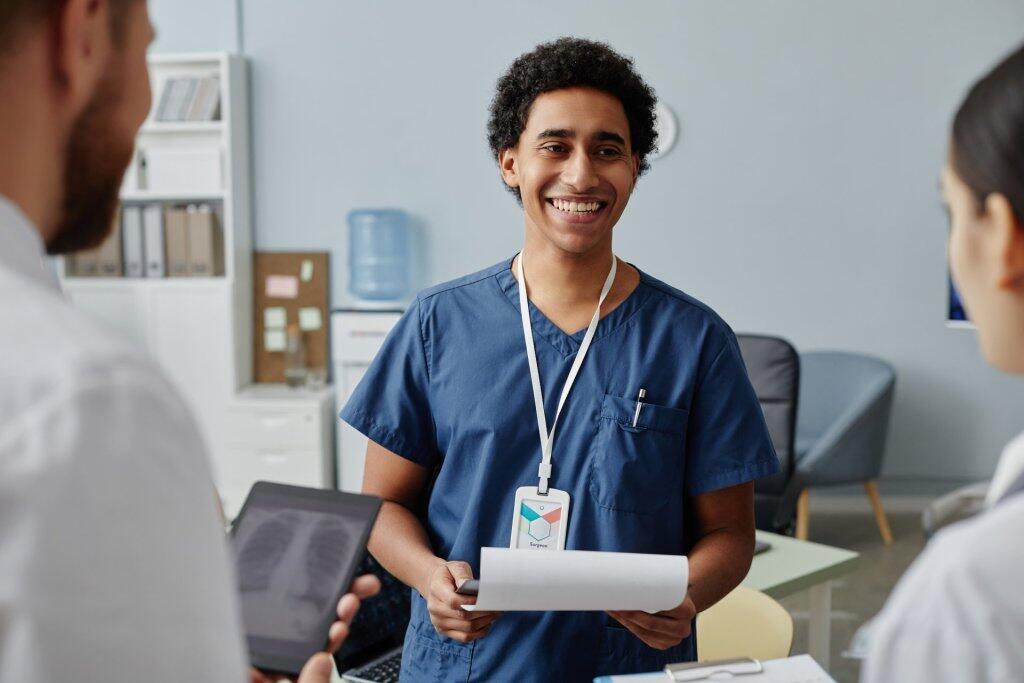 Smiling medical student in blue scrubs holding paperwork while talking with colleagues in a clinical setting