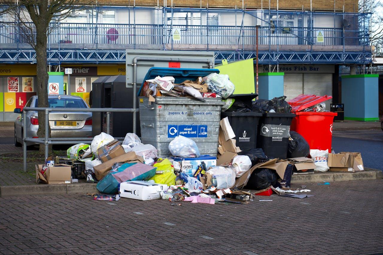 Overflowing dumpsters surrounded by scattered trash and cardboard, highlighting poor waste management in an urban area.