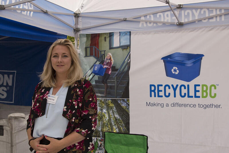 A woman stands at a booth with a RecycleBC banner, promoting recycling with visuals of people engaging with recycling in their community.