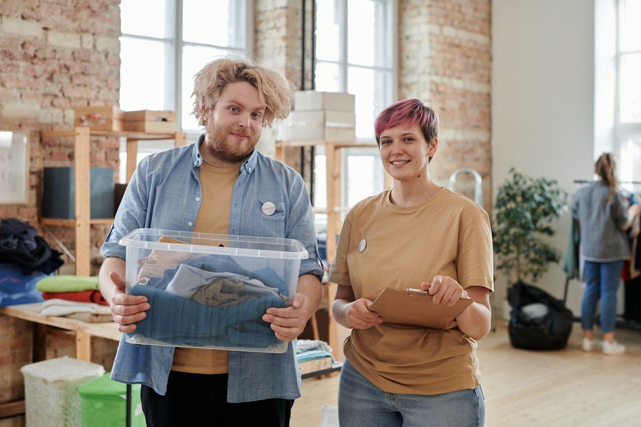 Two people in casual attire stand in a clothes donation center, one holding a box of clothes and the other with a clipboard.