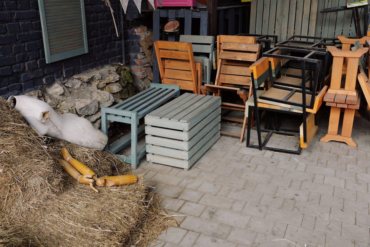 A cluttered outdoor space displaying stacked wooden chairs, tables, and a hay pile, with a white object partially hidden in the background.