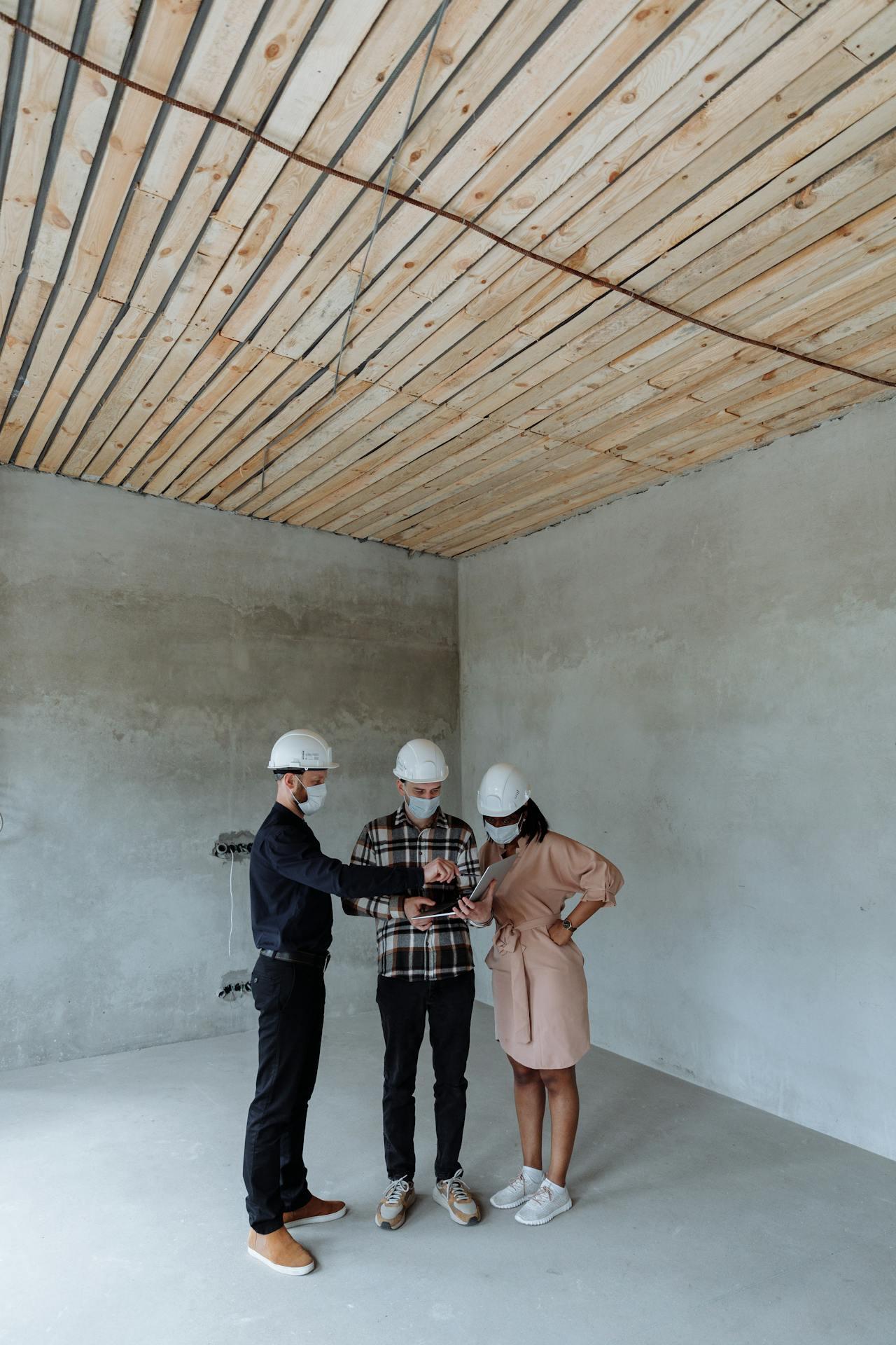Three individuals in construction helmets collaborate in an unfinished room with a wooden ceiling and gray walls, discussing plans on a tablet.