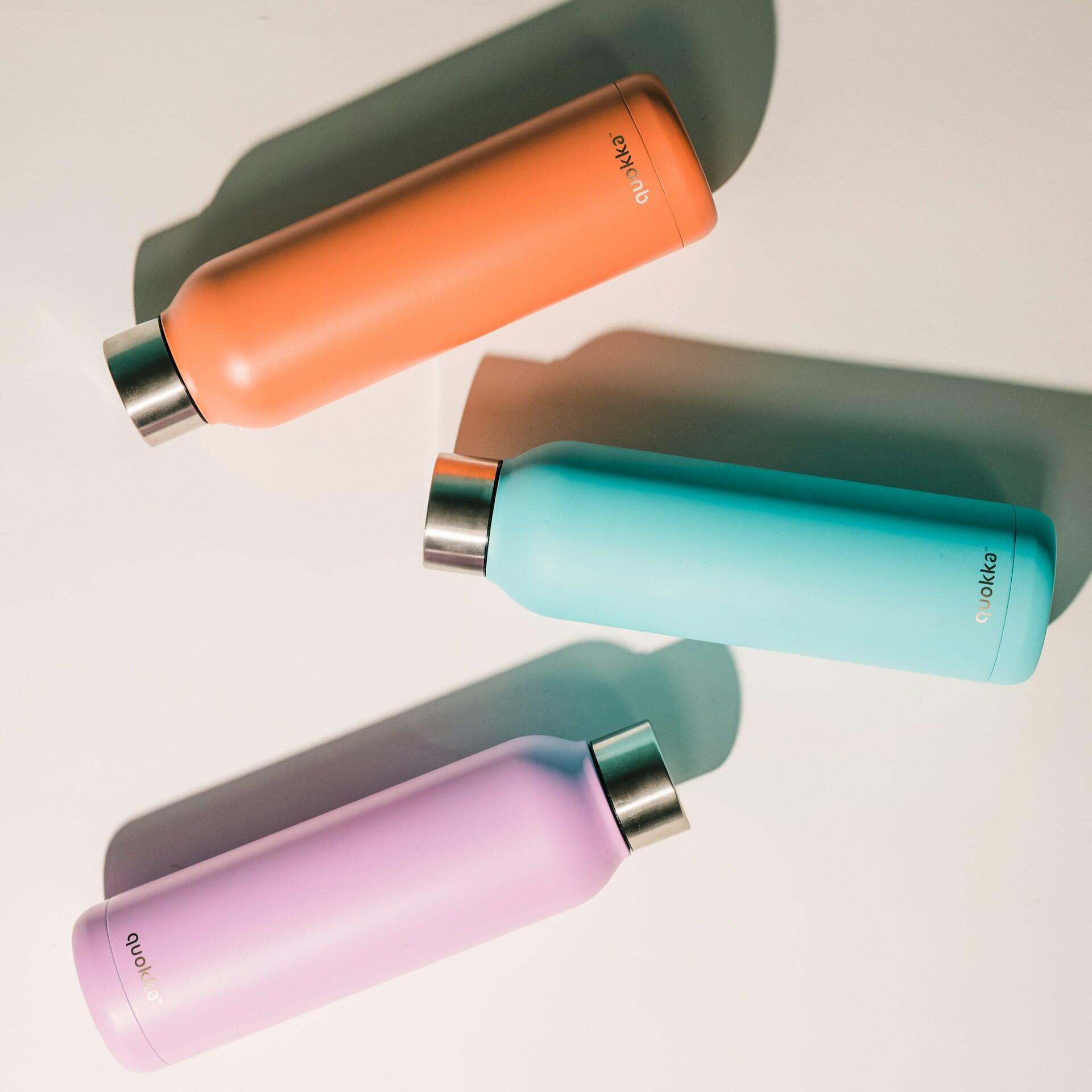 Three colorful stainless steel water bottles in orange, teal, and lavender, arranged on a light background.