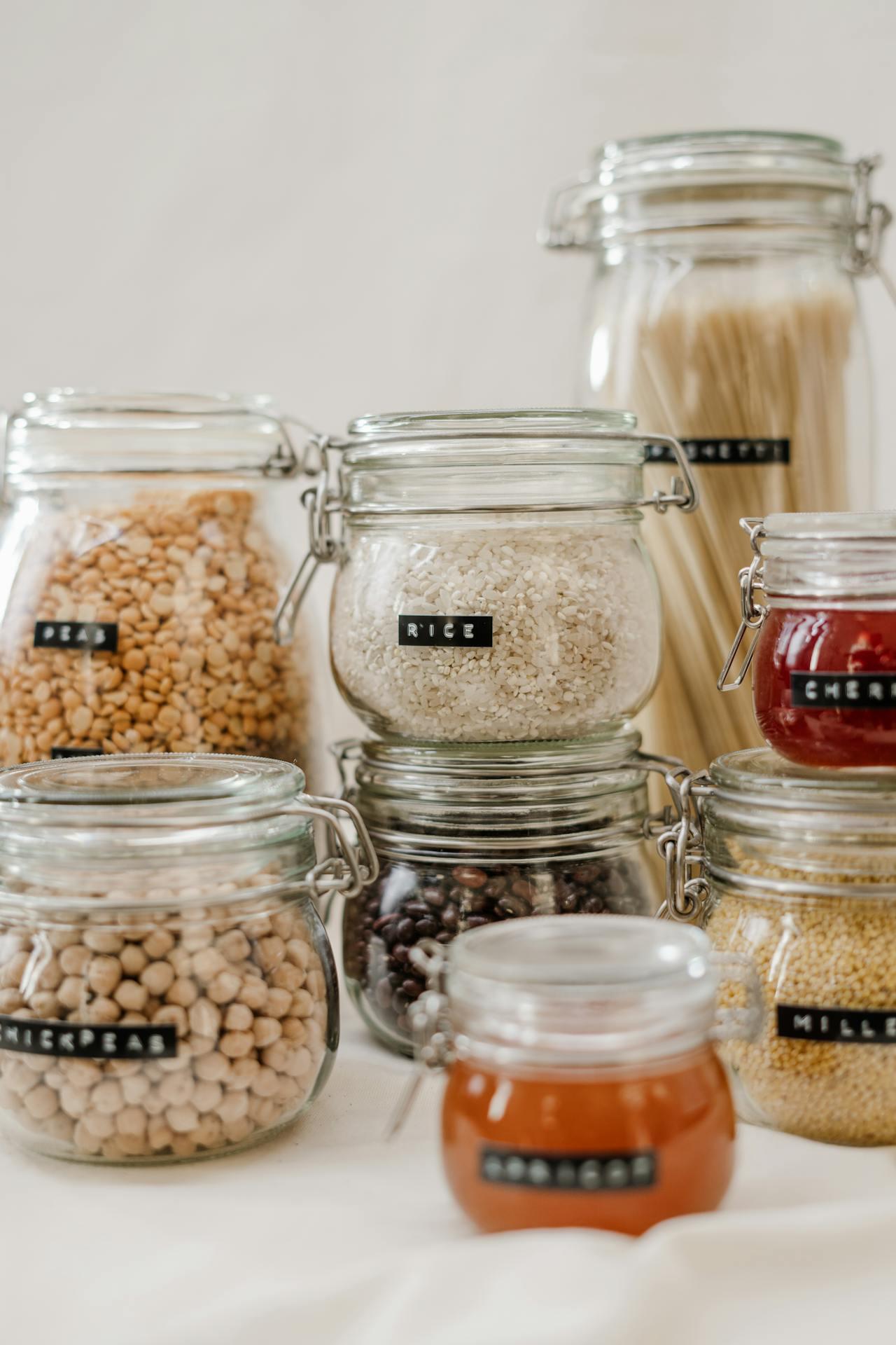 Glass jars filled with various grains, legumes, and preserves, labeled clearly, arranged aesthetically against a light background.