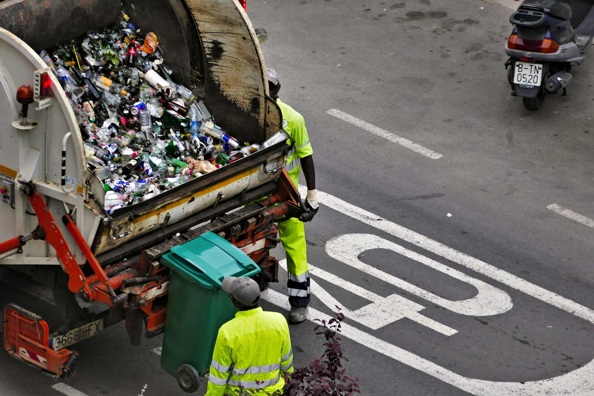 Two sanitation workers in neon yellow vests collect recyclables from a truck overflowing with bottles and cans on a city street.