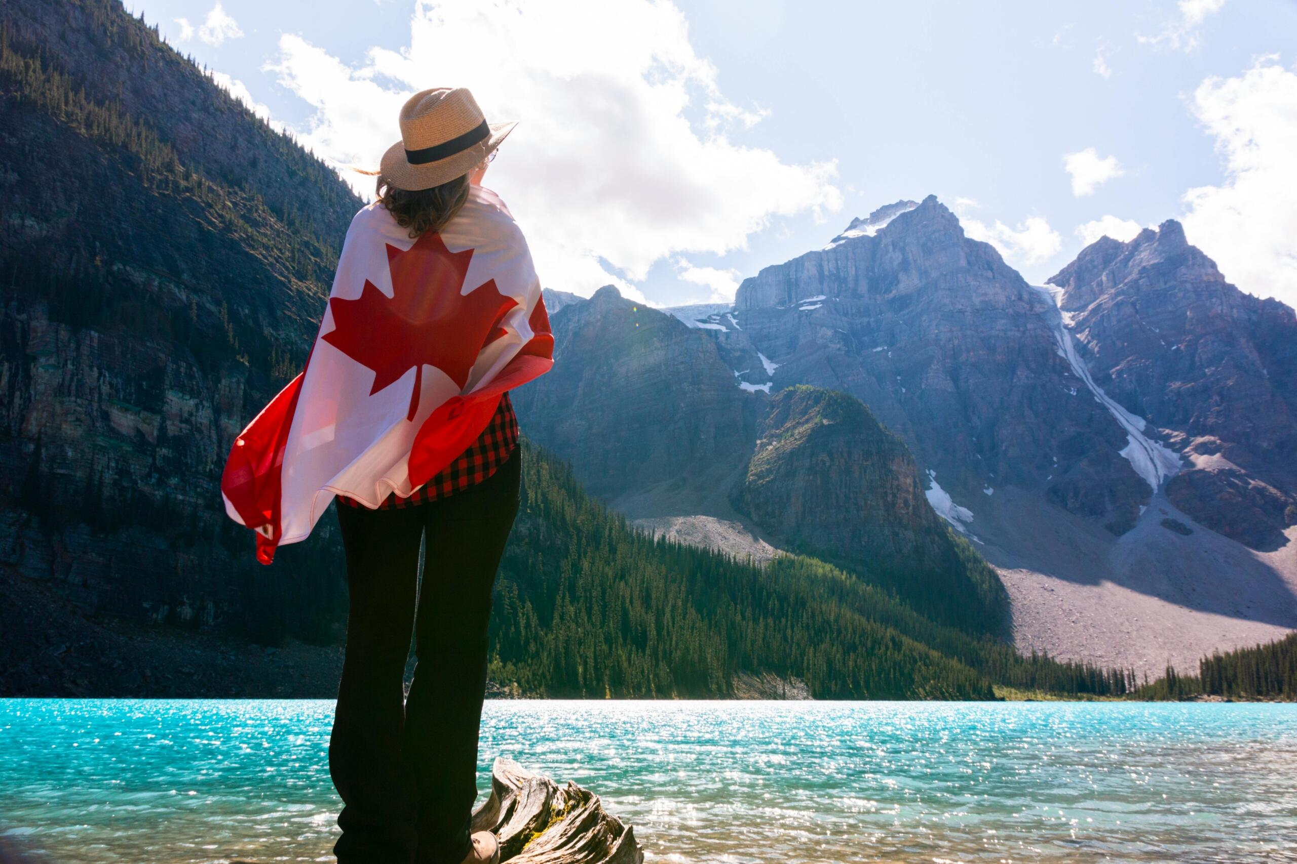 A woman wearing a Canadian flag on her back stands in front of a crystal clear blue lake, looking at the mountains.