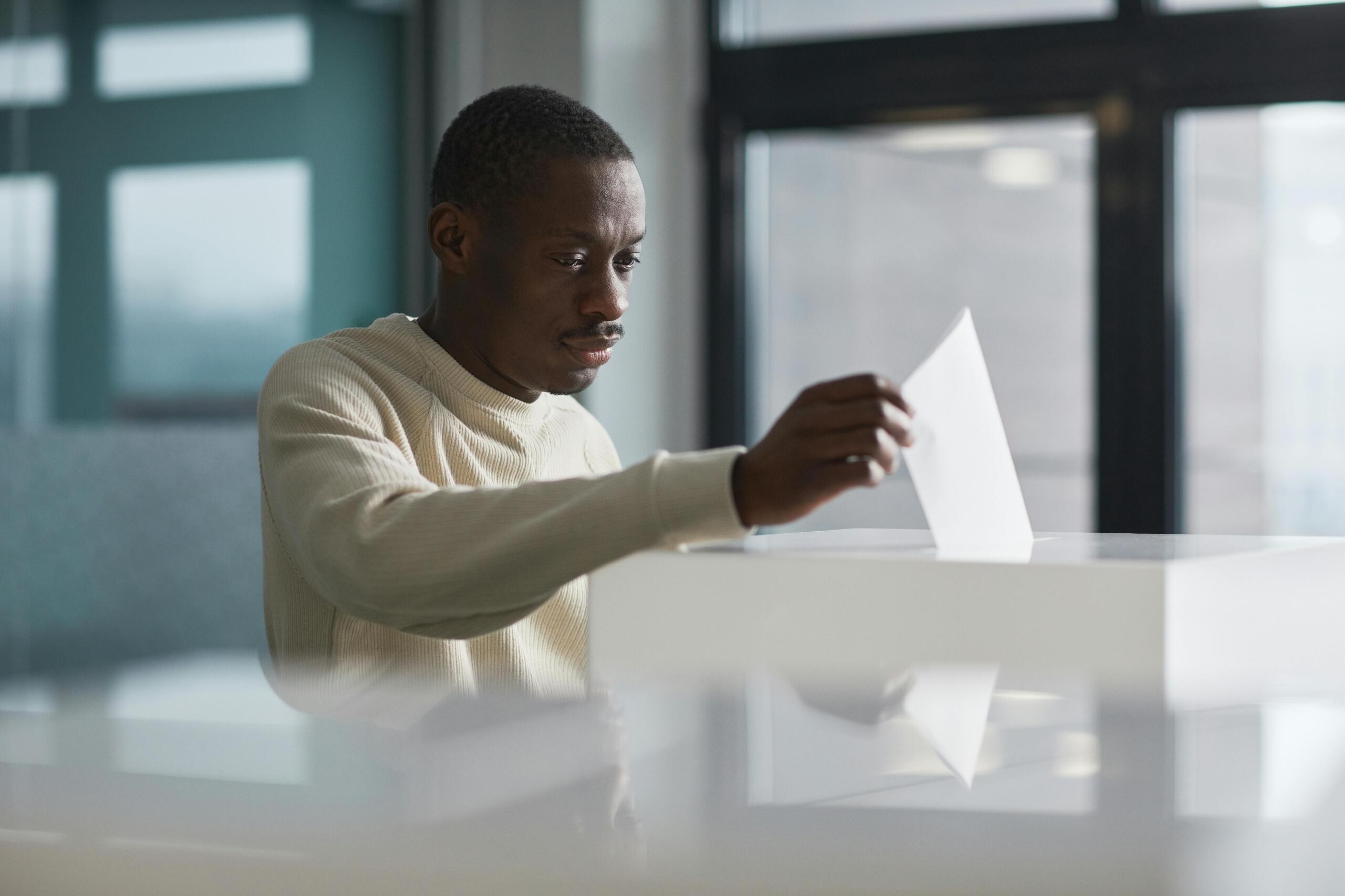 a man casts his vote into a ballot box, his face solemn.