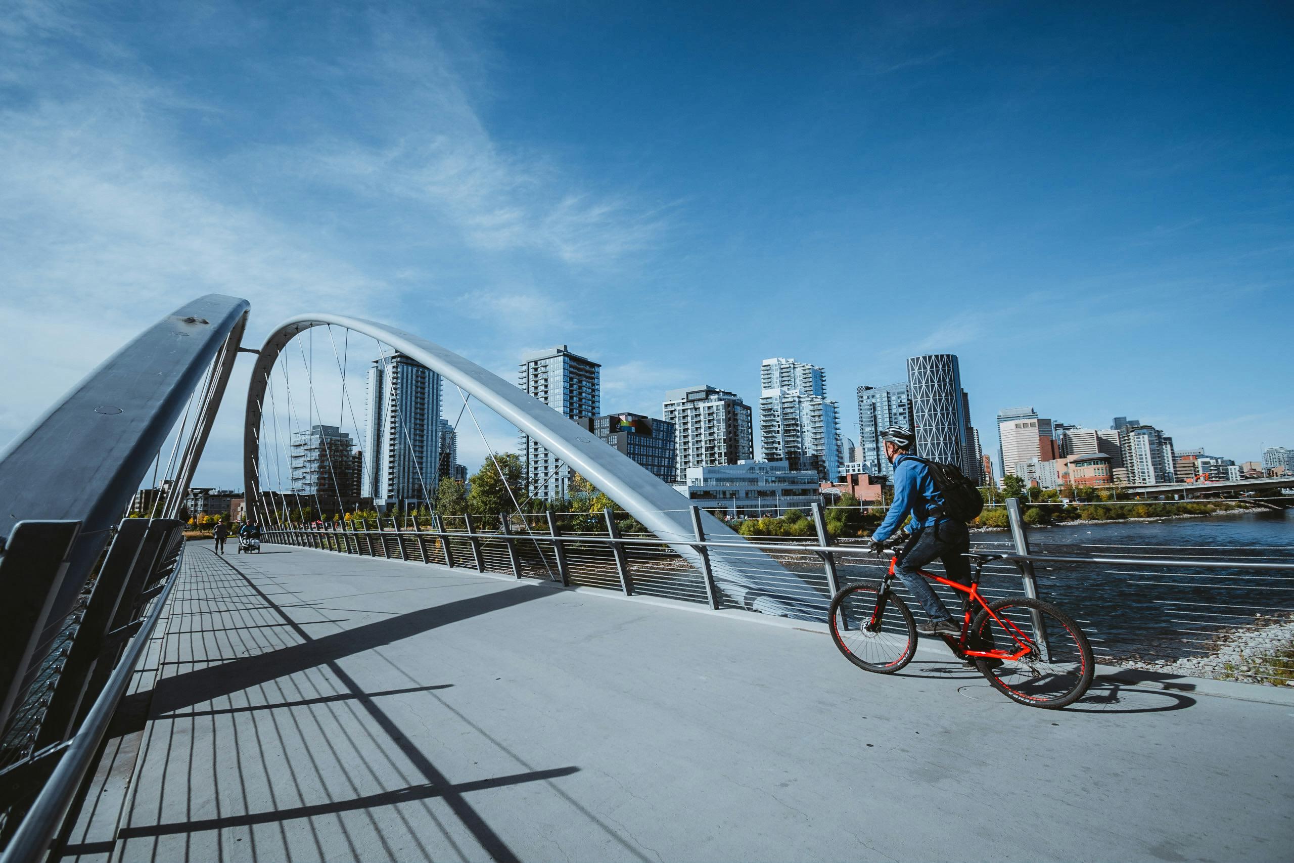 A photo of a man cycling over a bridge in Calgary, the skyline looming in the background. The day is clear and sunny.