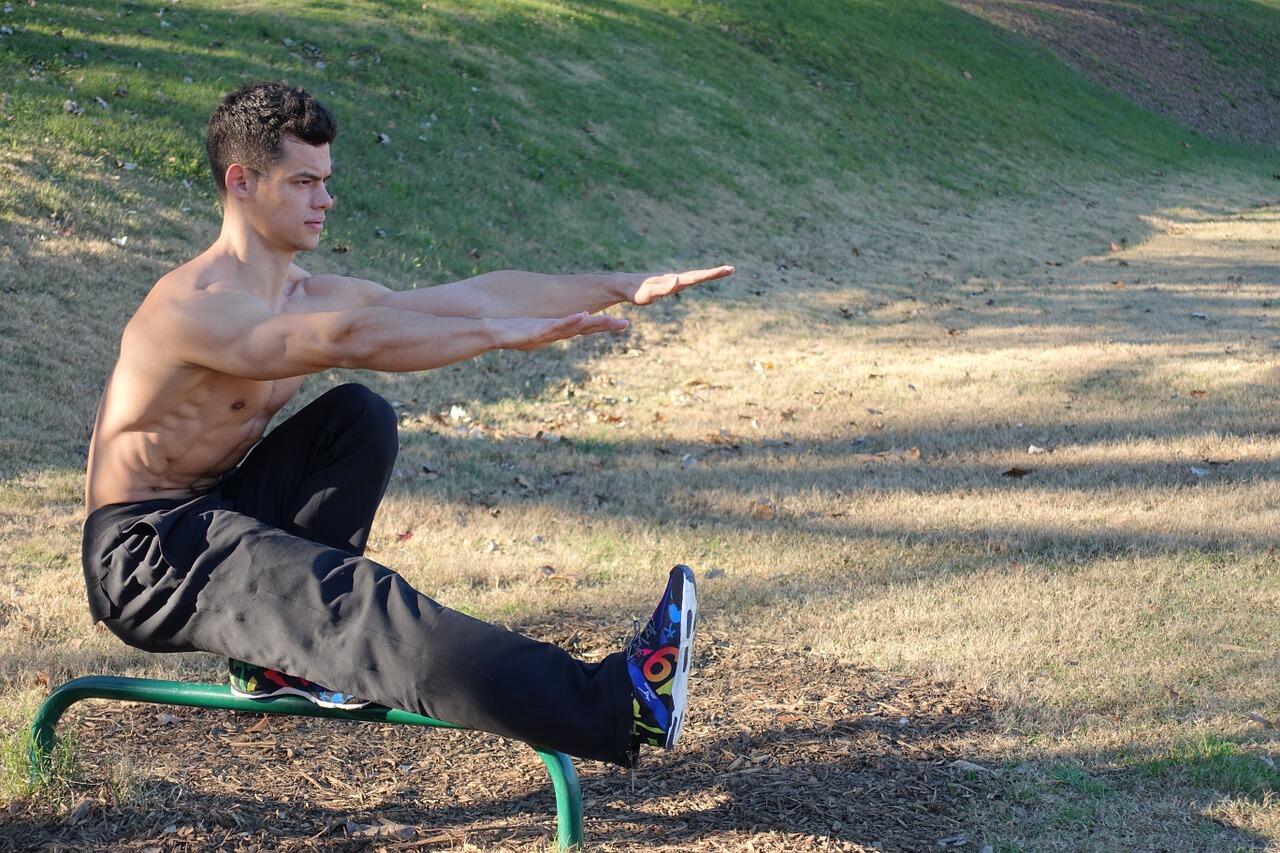 A shirtless man performing a pistol squat on an outdoor bench in a grassy park, demonstrating balance and leg strength as part of a calisthenics workout.