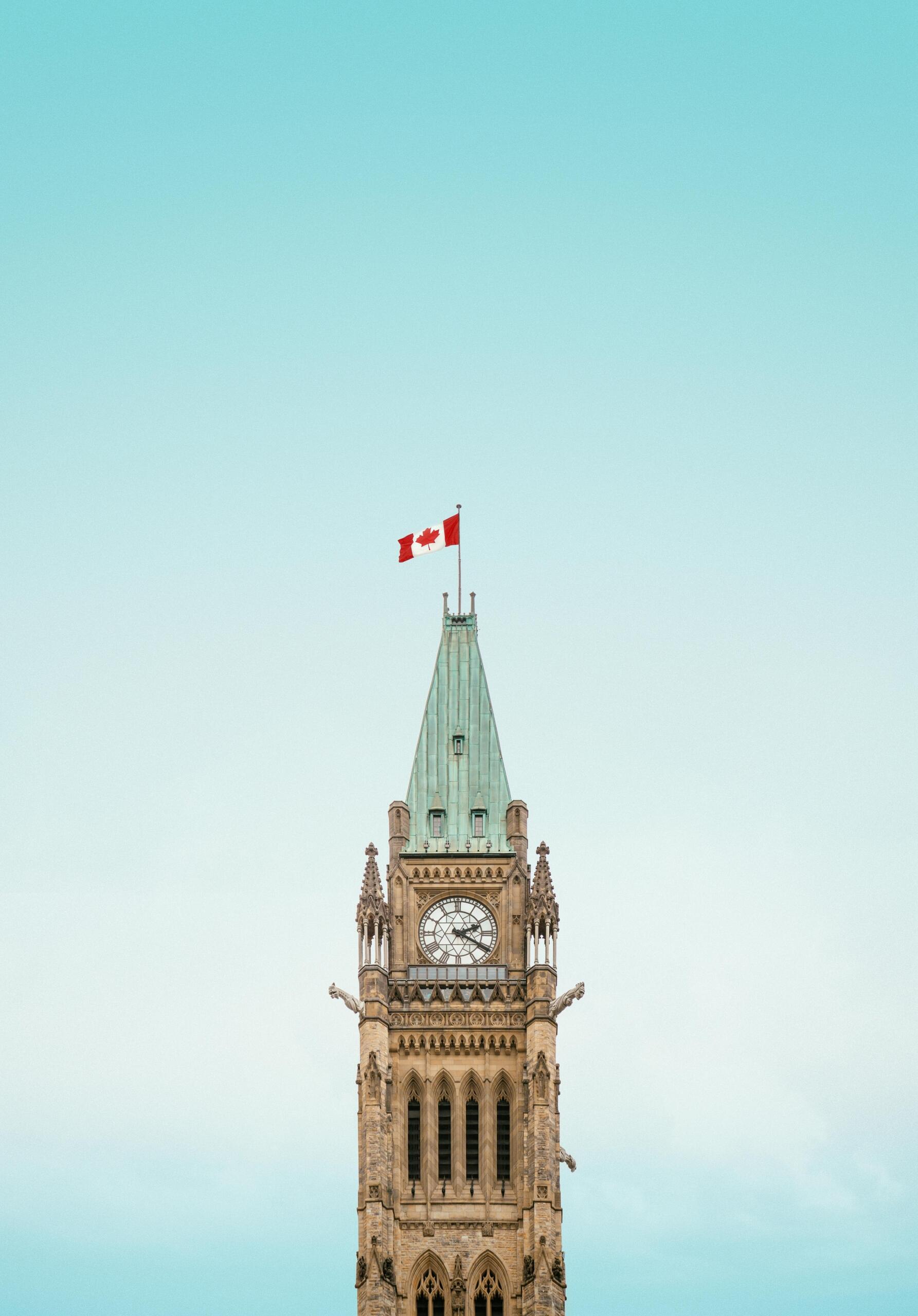The clock tower at Parliament Hill in Ottawa pictured in front of a clear sky, a Canada flag waving in the wind at the top.