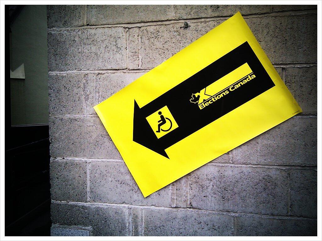 A bright yellow Elections Canada sign with a black arrow pointing downward, with the icon of a person in a wheelchair.