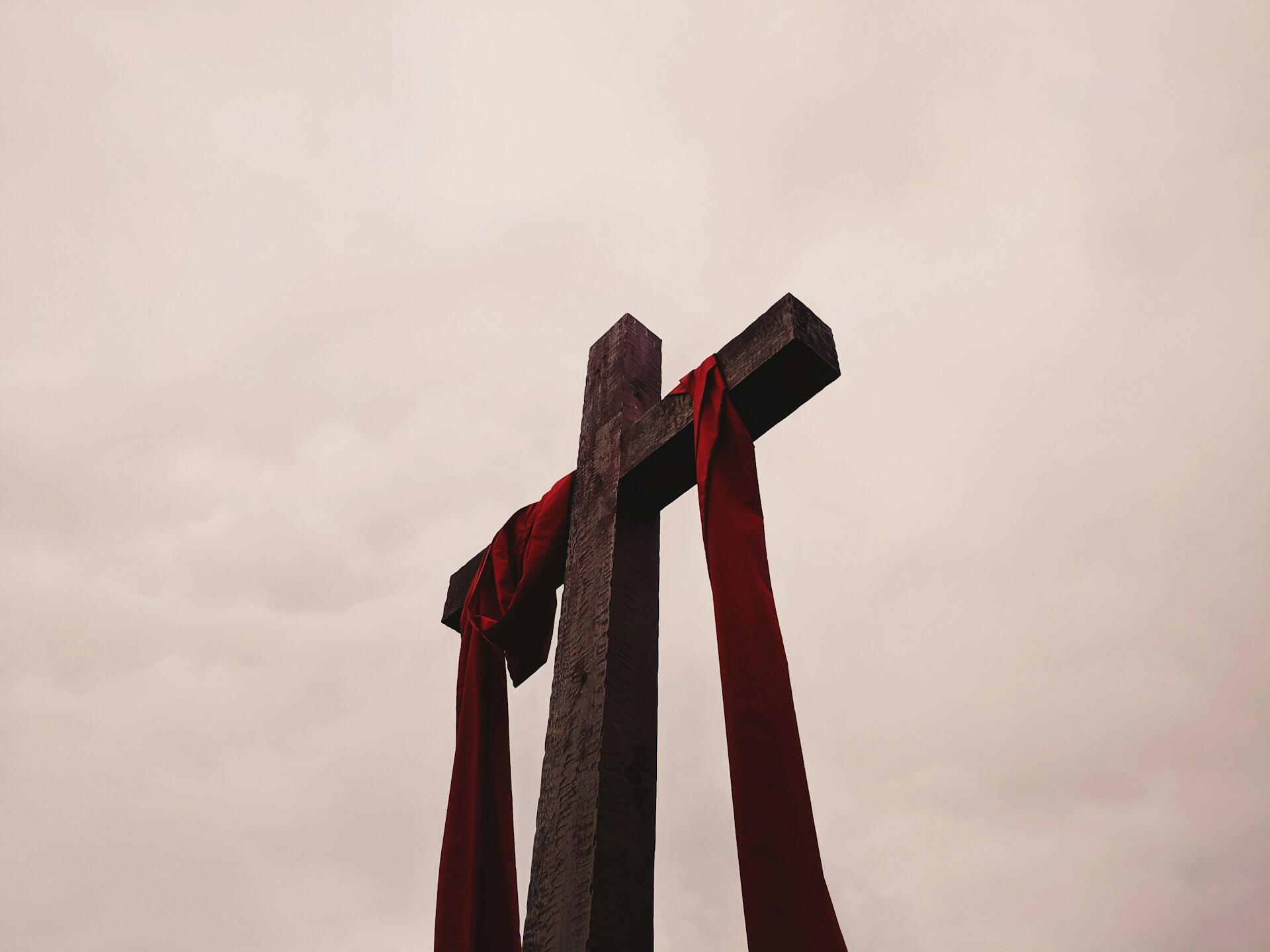 A wooden cross adorned with flowing red ribbons against a cloudy, muted sky, symbolizing faith and remembrance.