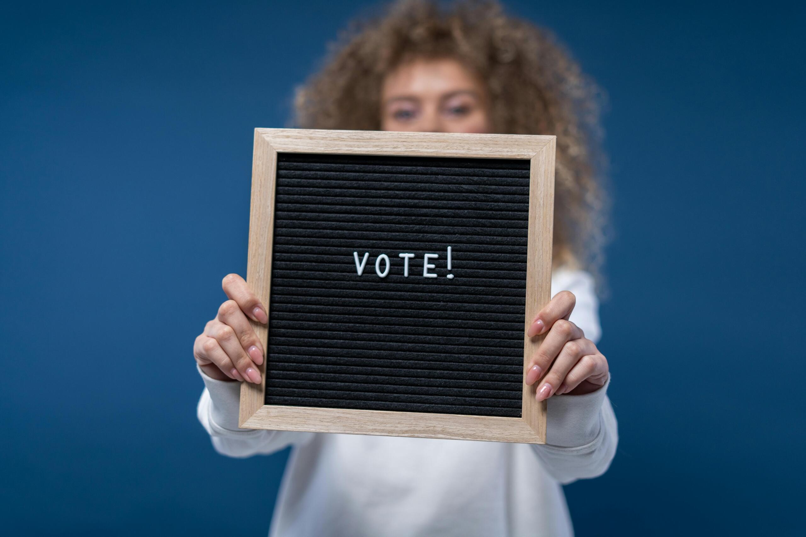 A woman holds a black and white letterboard saying the word "vote!"
