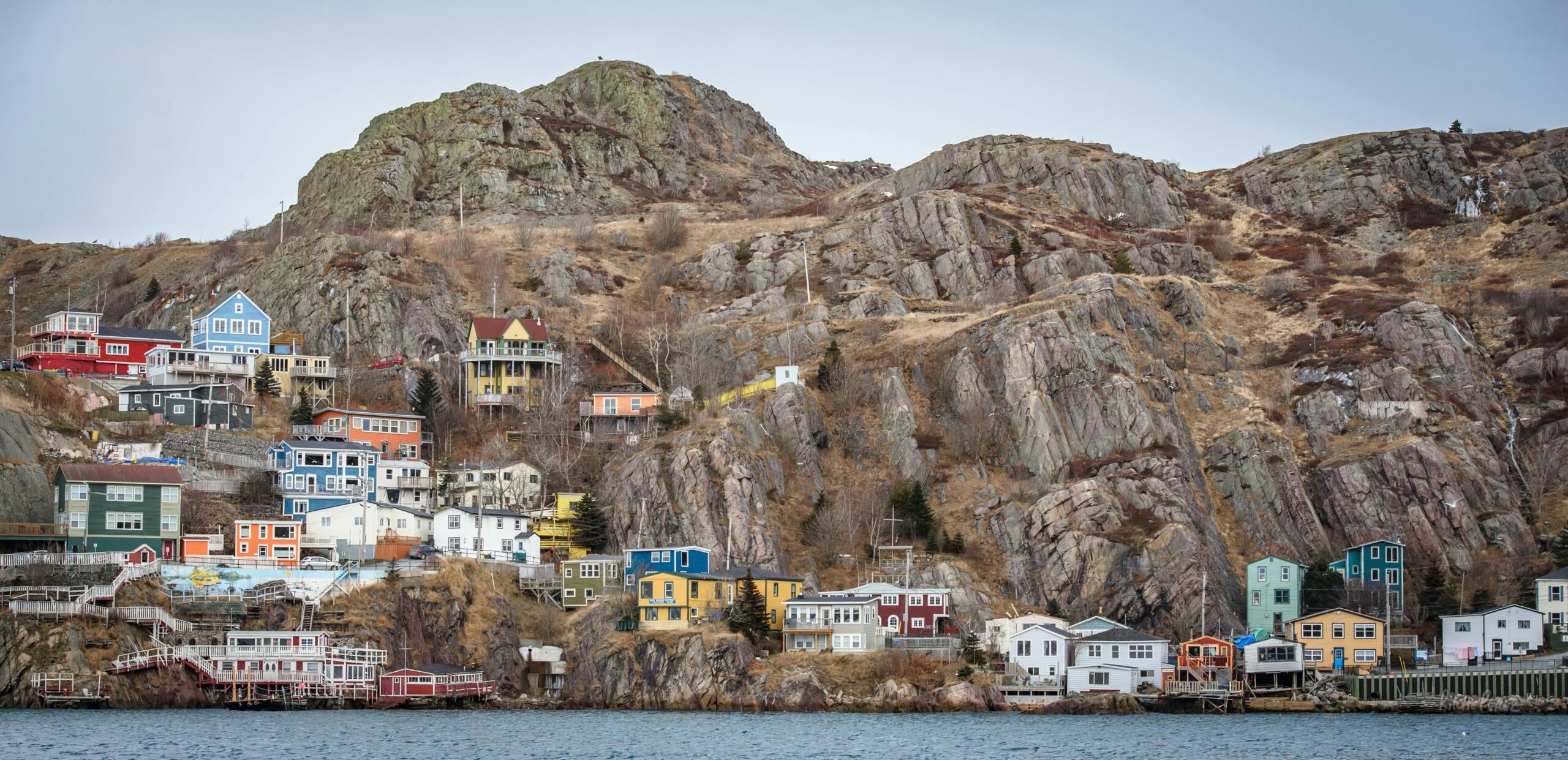 A small coastal town in Newfoundland with brightly painted wooden houses perched along the rocky shoreline, fishing boats anchored in the harbour, and rugged cliffs rising in the background under a cloudy sky.