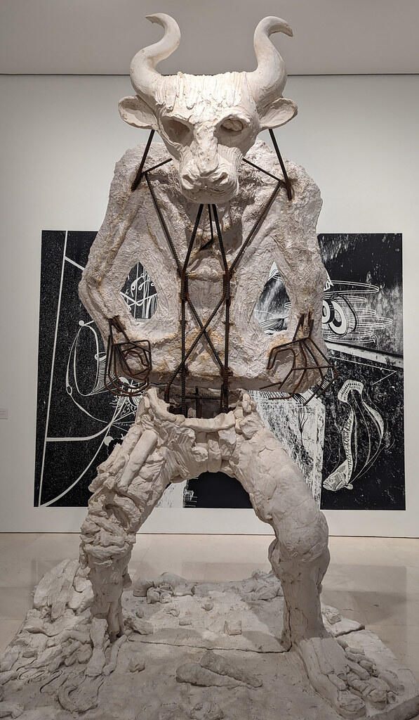 A large white sculpture of a bull-headed figure with exposed metal framework, set against a contrasting black and white art backdrop.