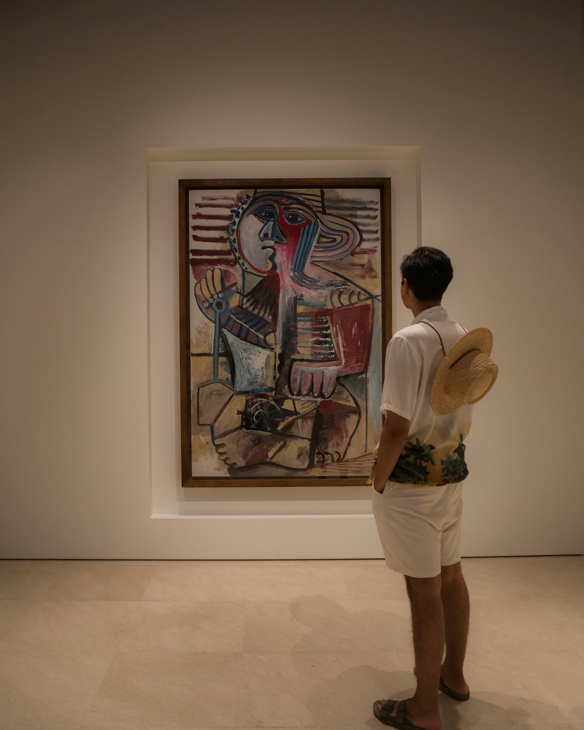 A person stands admiring a colorful Picasso painting in a minimalist gallery, framed by a soft, neutral background.