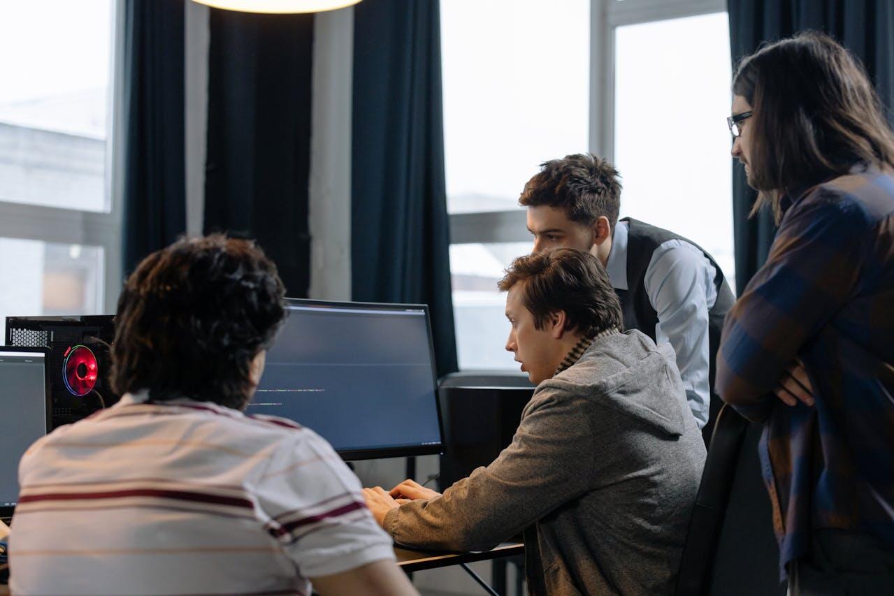 A group of four individuals collaborating at a computer, focused on programming or tech work in a modern office setting.