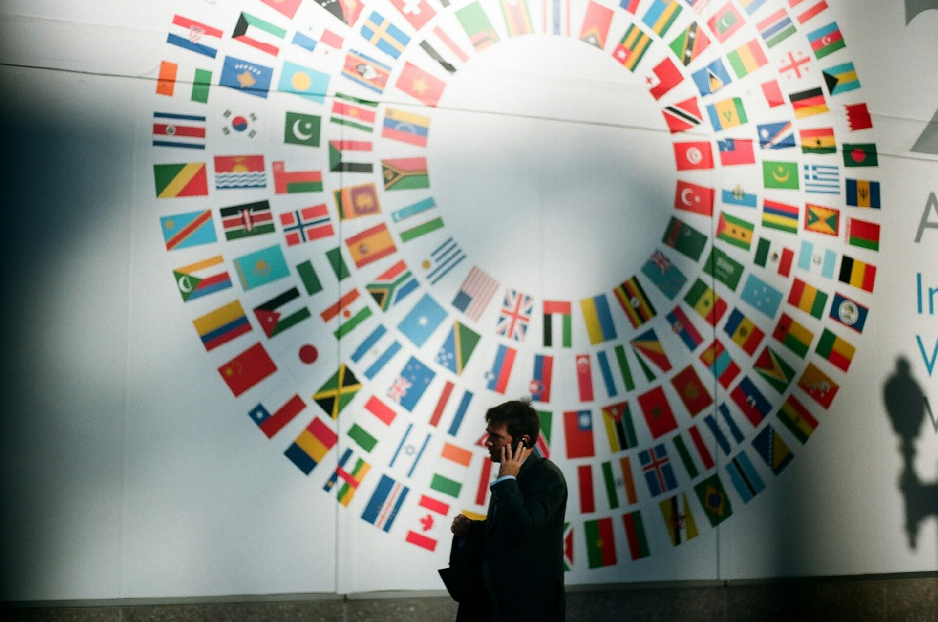 A man in a suit stands in front of a large mural featuring numerous national flags arranged in a circular pattern.