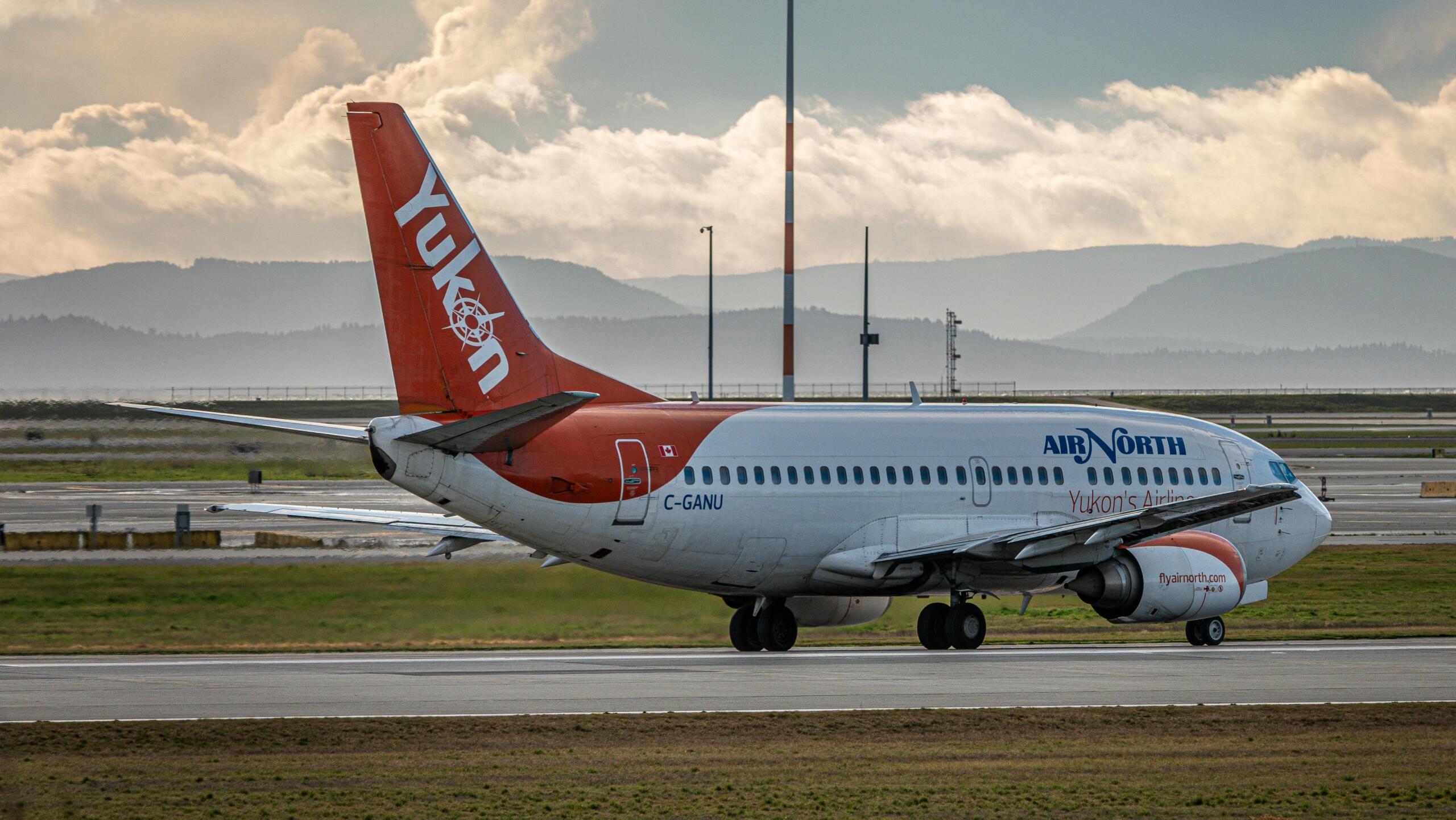 An "Air North" Plane with Yukon written on the tail stands on a runway at an airport. Rolling hills can be seen in the background.