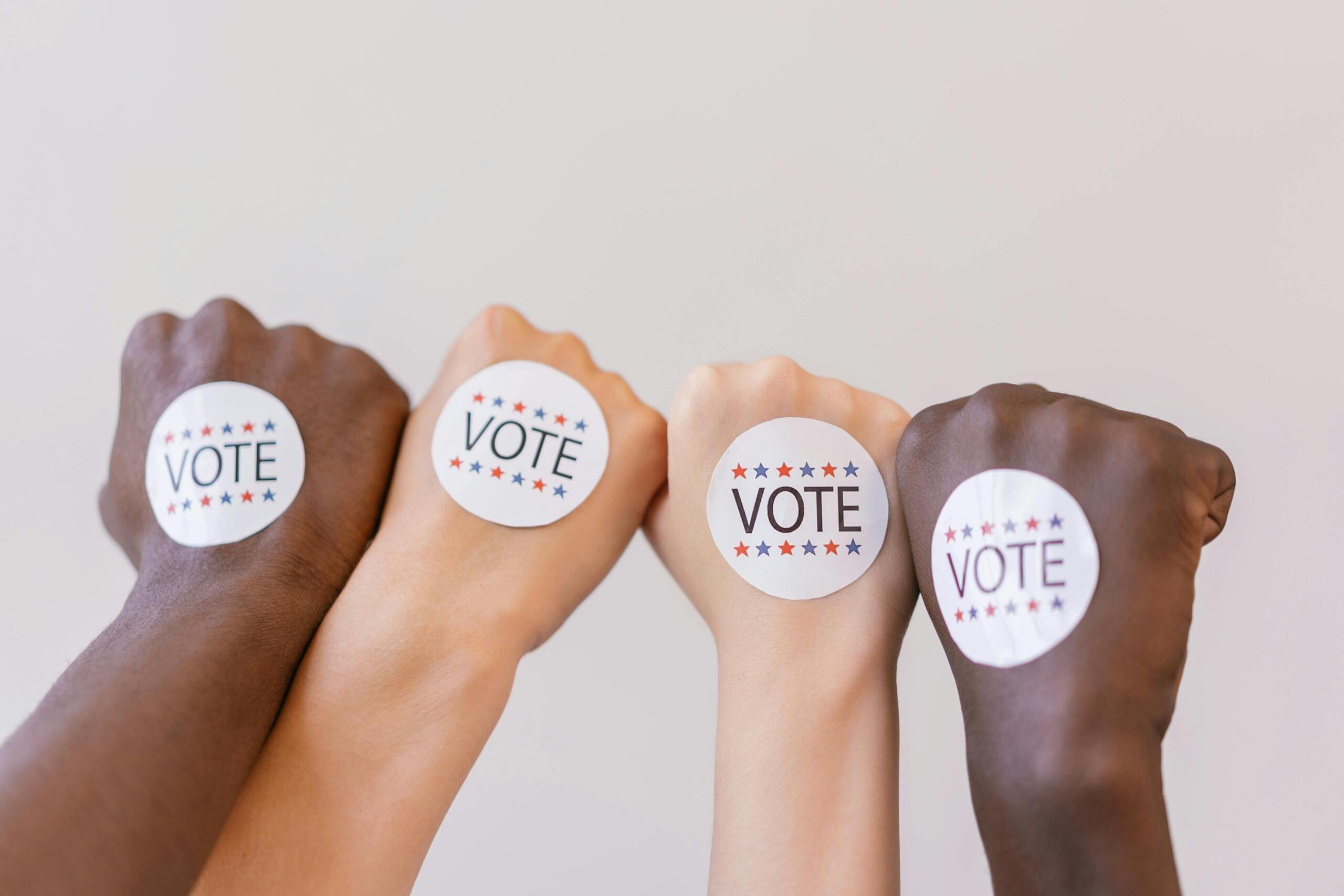 Multiple hands, reflecting the diversity of modern communities, make fists. A "vote" sticker is on each hand.