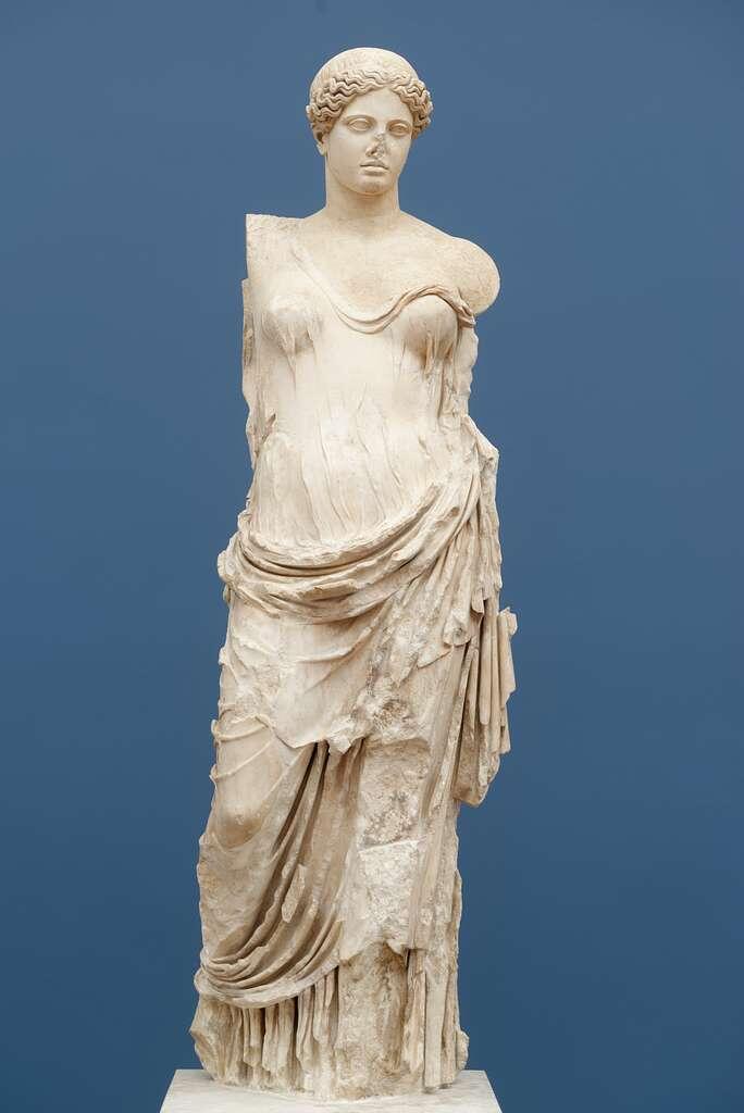 Ancient marble statue of a woman in draped clothing against a blue background. The figure is missing arms and displays a serene expression.