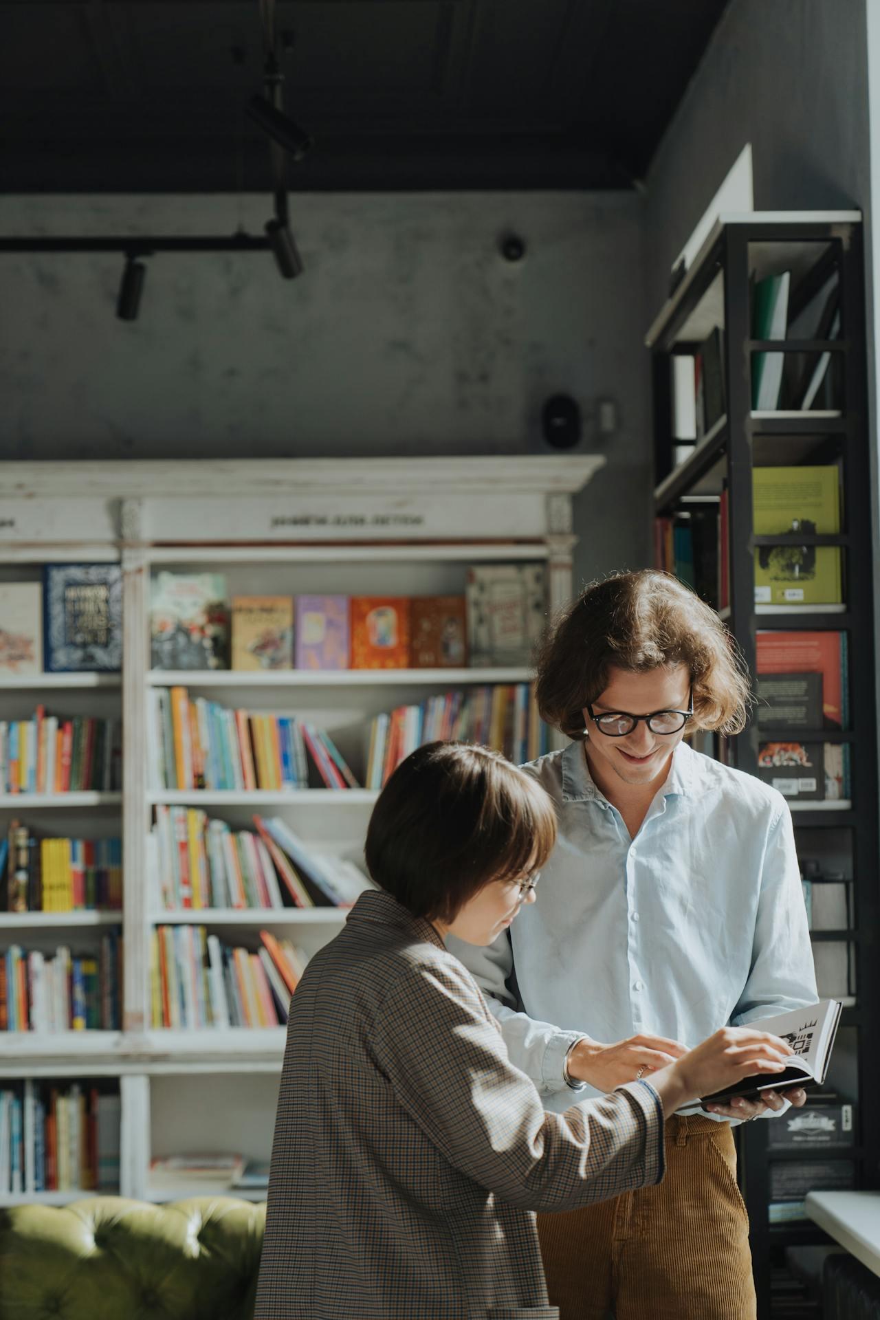 A woman in a brown checked jacket interacts with a man in a light blue shirt, both looking at a small book in a cozy library setting.