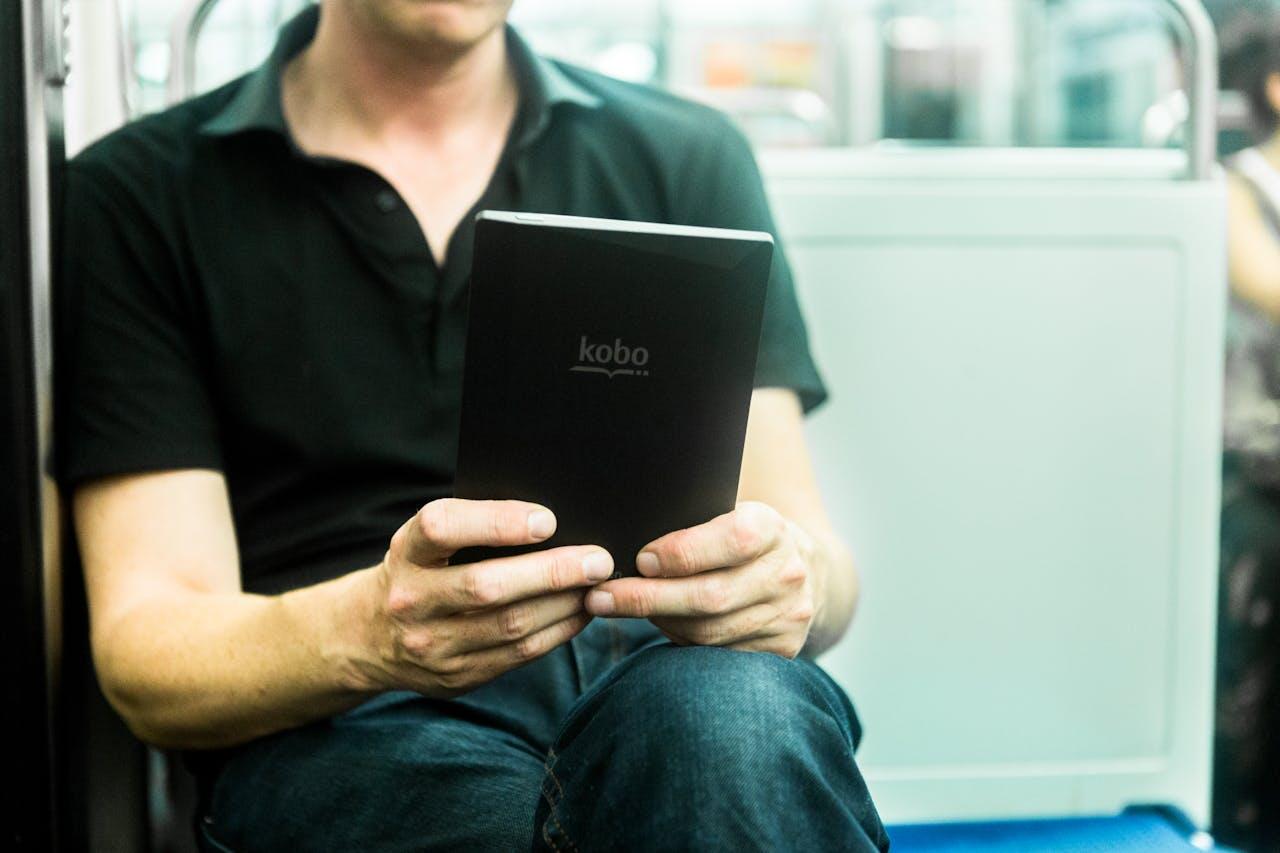 A person in a black polo shirt sits on a subway, reading a Kobo e-reader with focused attention.