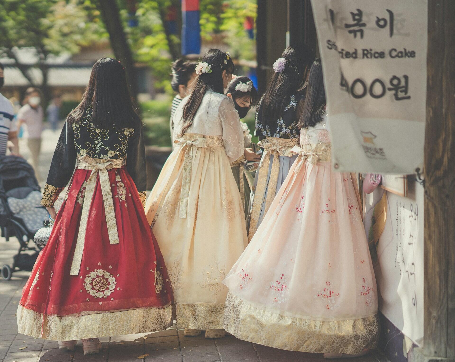 Four women in traditional hanbok attire, featuring vibrant colors and intricate designs, stroll past a market stall in a sunny setting.