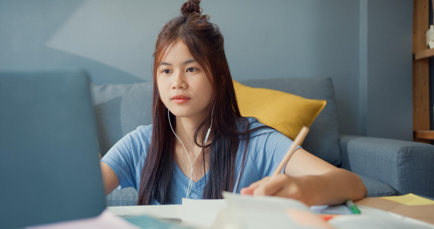 A person studying at a desk, wearing headphones, with a laptop and scattered notes in a cozy living room setting.