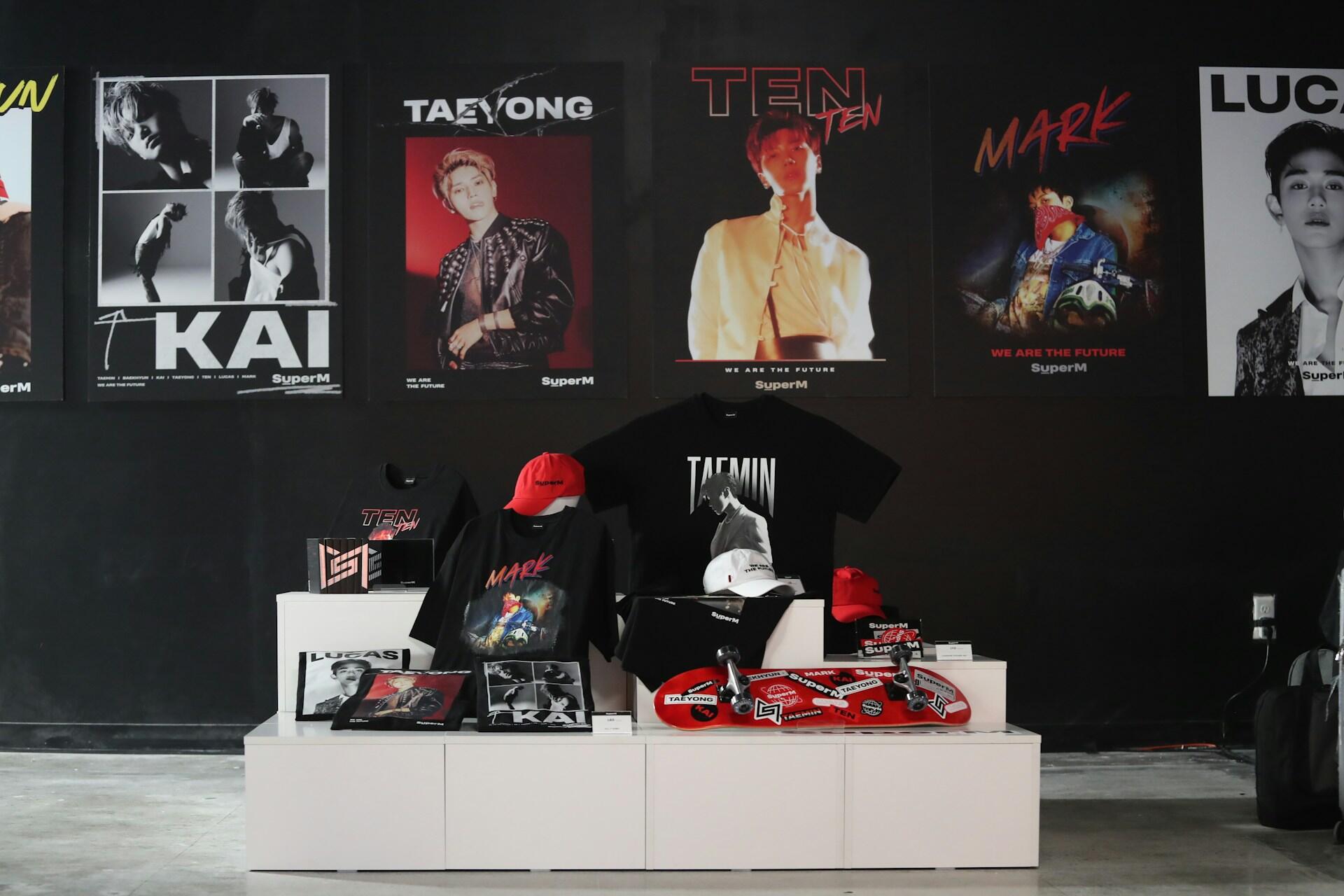 A display of SuperM merchandise including shirts and skateboards, and posters of various members against a dark background.