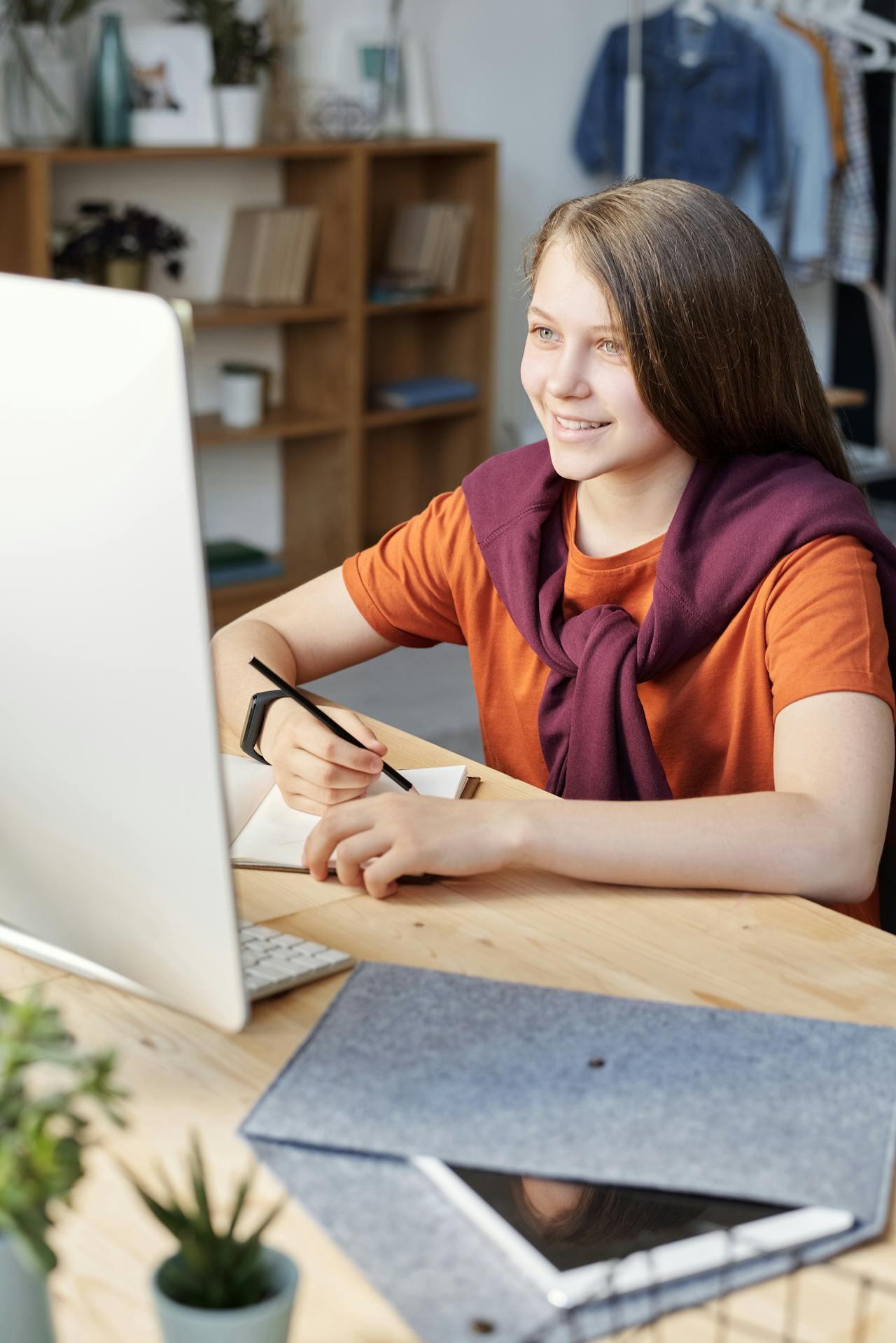 A person sits at a desk, writing in a notebook with a pencil, surrounded by plants, a computer, and office supplies.