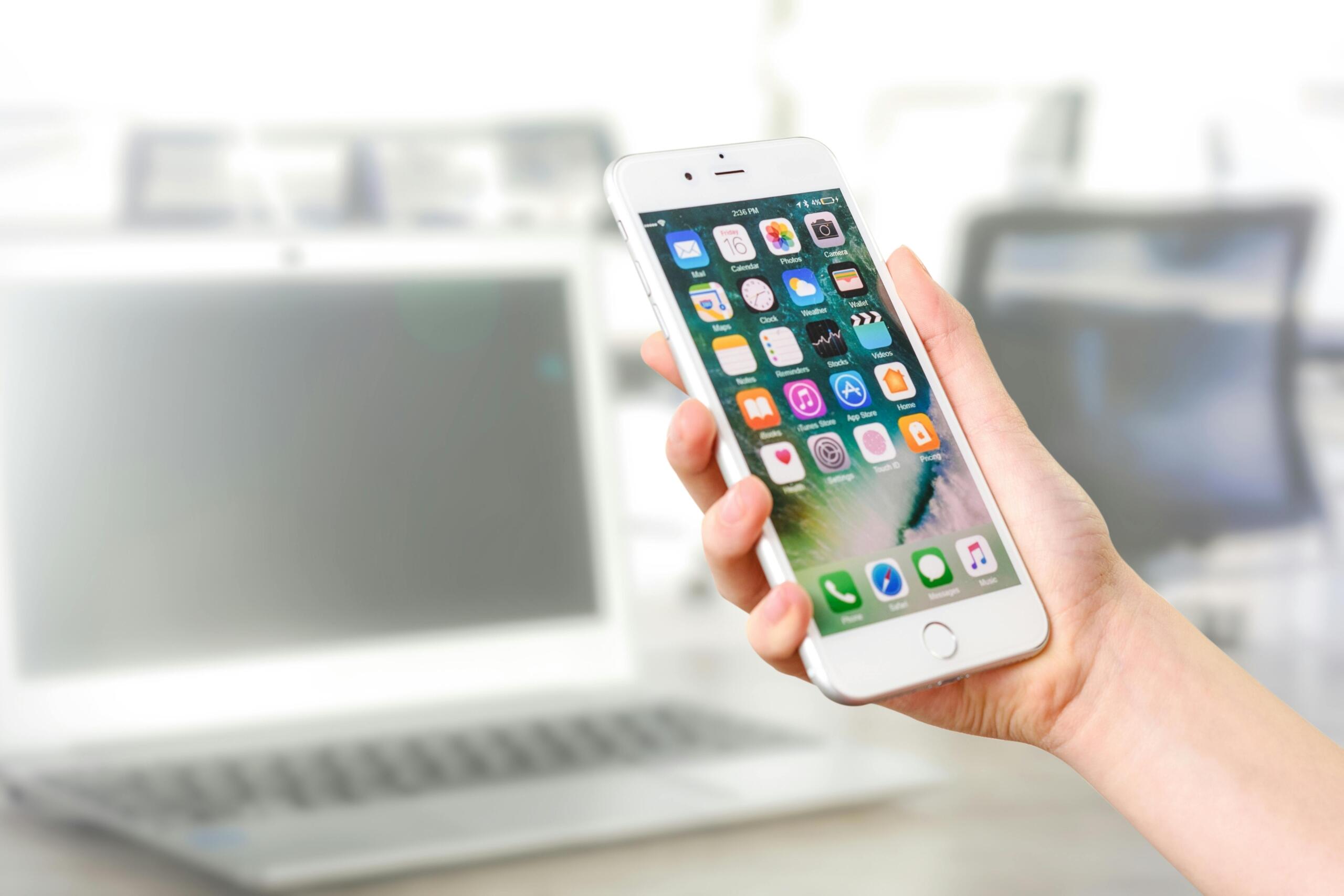 A person holding a smartphone displaying various app icons, with a blurred laptop in the background.